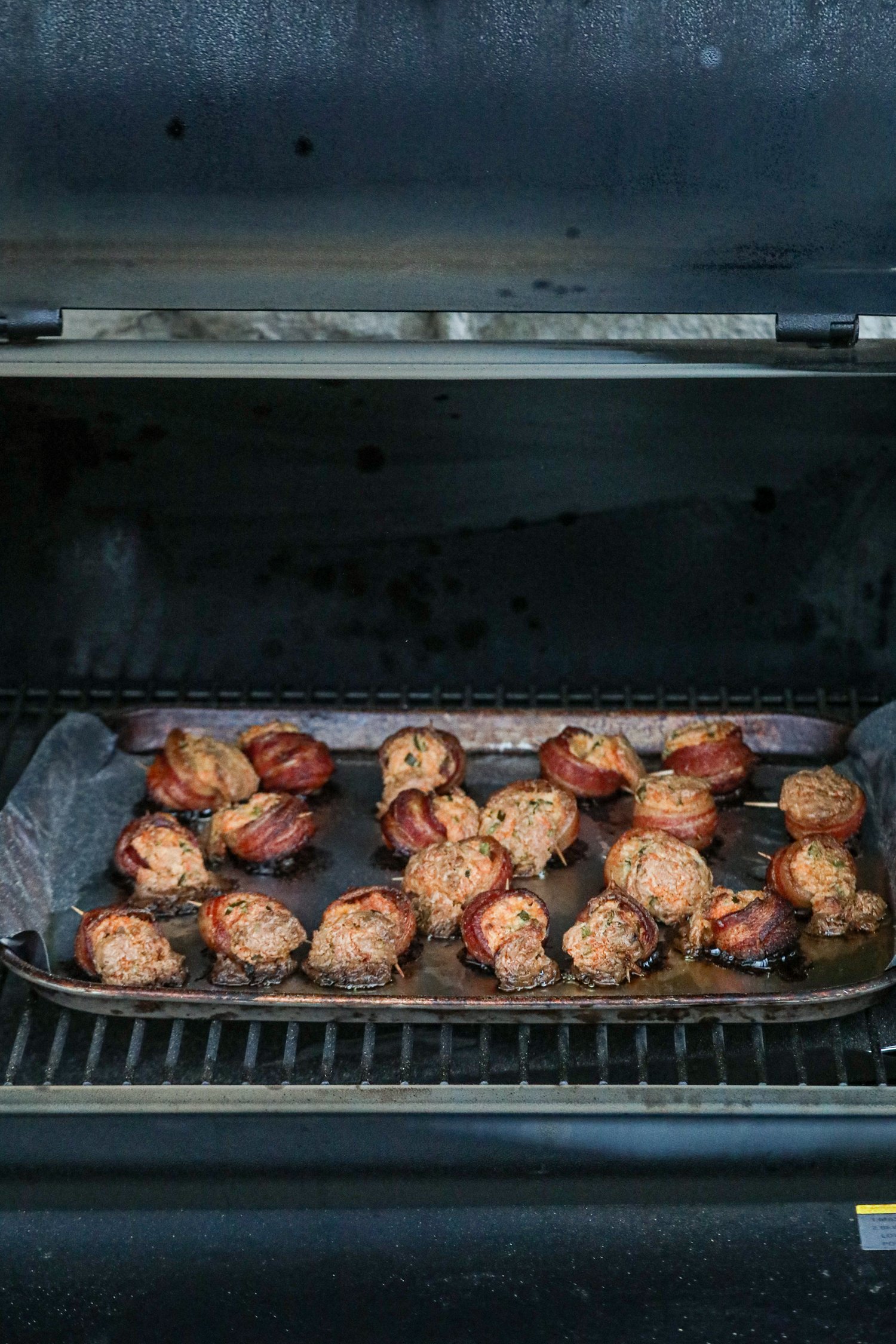 A tray of smoked pig shots inside a smoker, fully cooked to a golden-brown color. The bacon-wrapped sausage rounds are crisp, and the filling appears slightly caramelized. The smoker lid is open, revealing the finished appetizer resting on a foil-lined baking sheet.