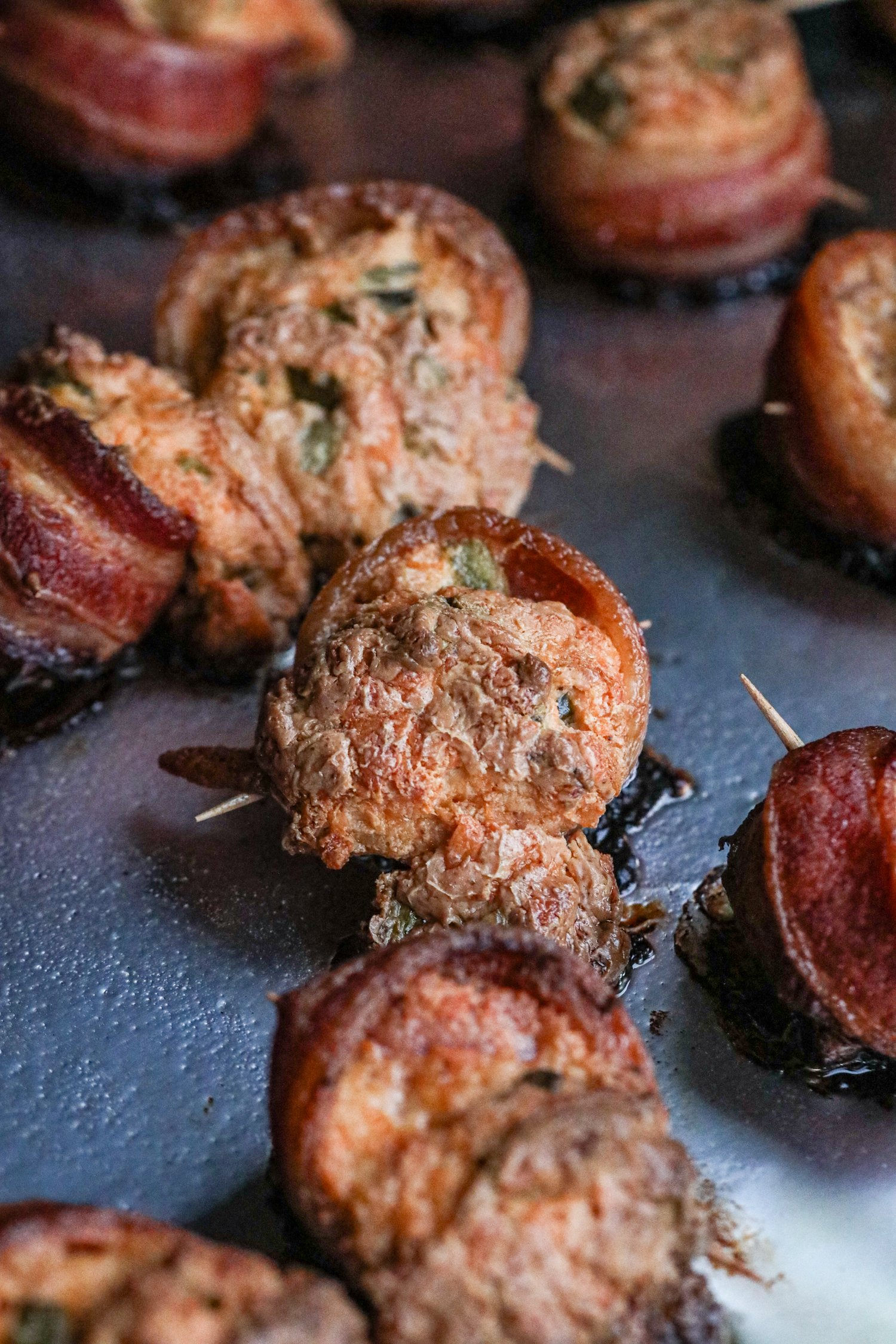 Close-up of smoked pig shots on a baking sheet, showing their crispy bacon-wrapped exterior and golden-brown filling. The filling has slightly puffed and caramelized, with bits of green herbs visible. Some of the appetizers have slightly crisp edges, highlighting their smoky, savory texture.