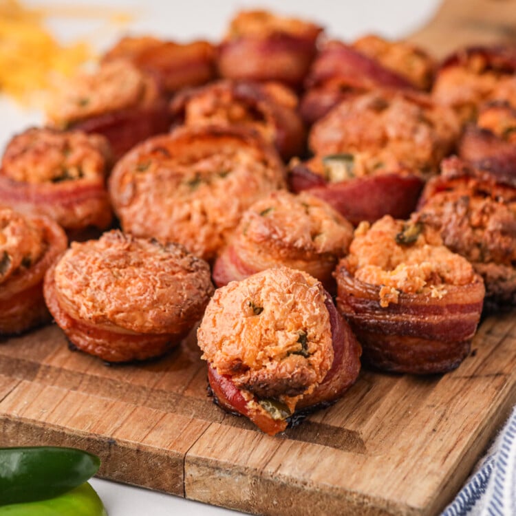 A wooden cutting board filled with freshly smoked pig shots. These bite-sized appetizers consist of sausage bases wrapped in crispy bacon and topped with a golden, cheesy filling with flecks of jalapeños. The pig shots have a slightly charred exterior, and the creamy filling has puffed up during cooking. In the background, shredded cheese and jalapeños are visible, adding to the vibrant and savory presentation.