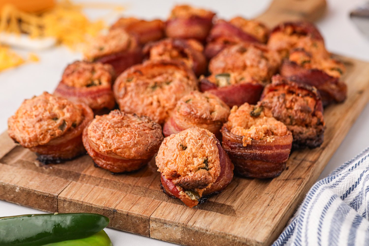 A wooden cutting board filled with freshly smoked pig shots. These bite-sized appetizers consist of sausage bases wrapped in crispy bacon and topped with a golden, cheesy filling with flecks of jalapeños. The pig shots have a slightly charred exterior, and the creamy filling has puffed up during cooking. In the background, shredded cheese and jalapeños are visible, adding to the vibrant and savory presentation.