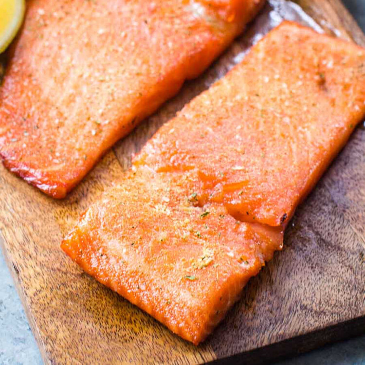Close-up of two fillets of smoked salmon on a wooden cutting board. The salmon has a rich, orange color with a slightly glossy texture, lightly seasoned with spices. A lemon wedge is partially visible in the background, adding a fresh contrast to the dish. The presentation emphasizes the flaky and tender nature of the smoked salmon.