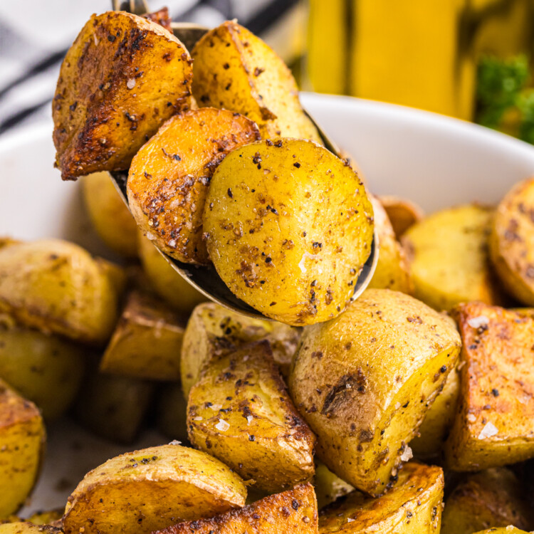 A close-up of crispy, golden-brown fried potatoes being scooped from a white bowl with a spoon. The potatoes are well-seasoned with black pepper, salt, and spices, giving them a flavorful crust. The texture is highlighted, showing the crispy edges and soft interior. In the blurred background, a bottle of olive oil, fresh parsley, and a black-and-white checkered napkin add to the inviting presentation.
