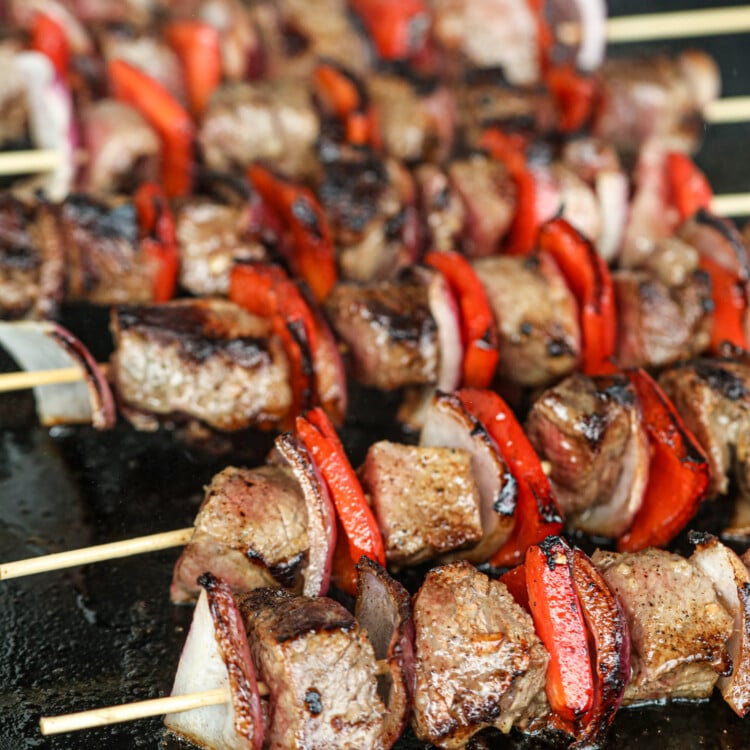 Close-up of grilled steak kabobs on a black cooking surface, featuring seared beef chunks, red bell peppers, and red onions. The skewers have a rich, caramelized crust, with visible grill marks and sizzling juices enhancing their glossy appearance.