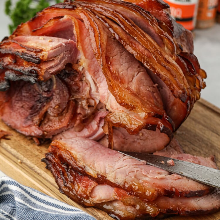 A close-up of a spiral-cut ham with a caramelized, crispy glaze on the outer layer, sitting on a wooden cutting board. A knife is partially slicing through the tender, juicy pink meat. In the background, spice jars and a salt and pepper shaker are visible on a kitchen countertop, with a striped kitchen towel in the foreground.