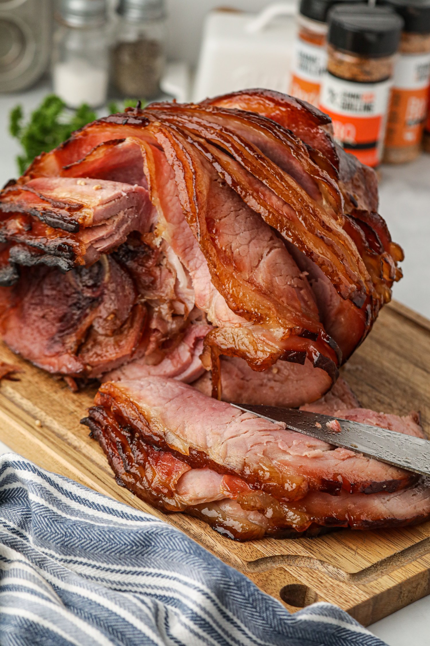 A close-up of a spiral-cut ham with a caramelized, crispy glaze on the outer layer, sitting on a wooden cutting board. A knife is partially slicing through the tender, juicy pink meat. In the background, spice jars and a salt and pepper shaker are visible on a kitchen countertop, with a striped kitchen towel in the foreground.