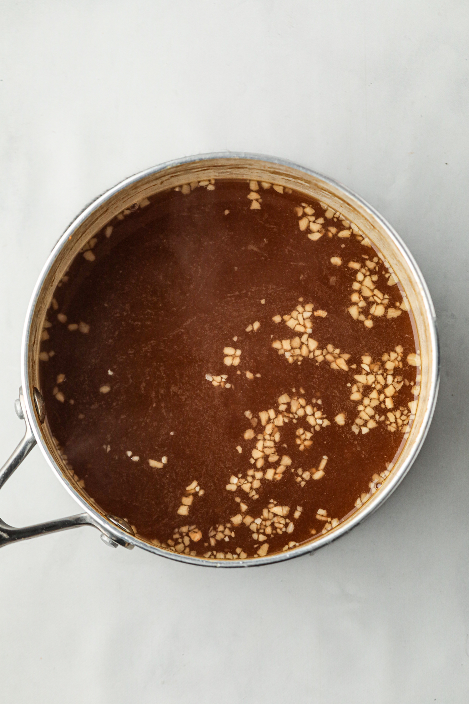 A stainless steel saucepan filled with a dark brown liquid, possibly a simmering sauce or broth, with finely chopped garlic pieces floating on the surface. The pot is placed on a light-colored background, and steam is faintly visible, indicating that the liquid is warm or cooking.
