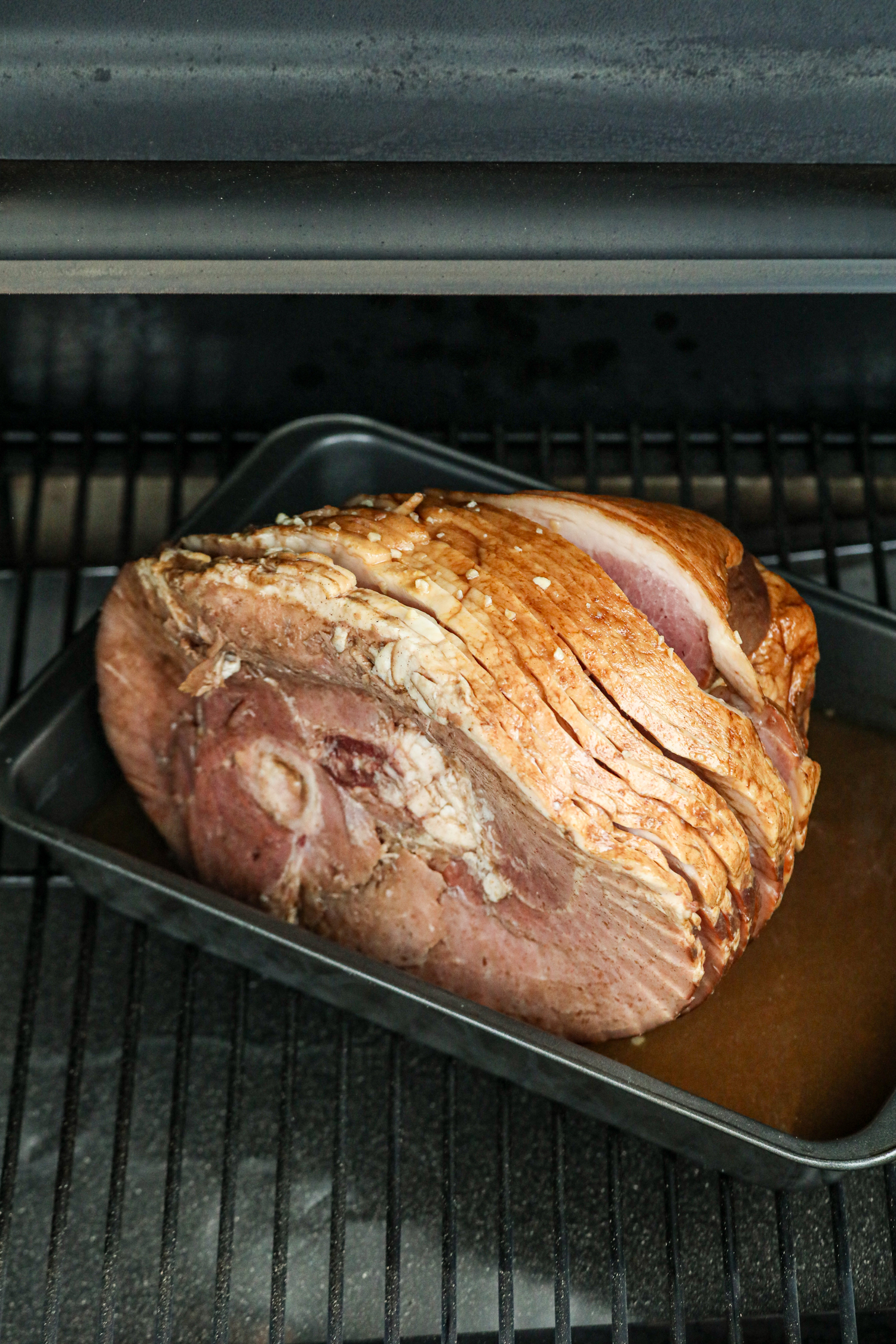 The spiral-cut ham has been placed on a grill inside a dark roasting pan filled with marinade or glaze. The ham is coated with seasonings, including minced garlic, and is beginning the slow cooking process. The grill grates are visible beneath the pan, and the smoky environment suggests it will develop a rich, flavorful crust.