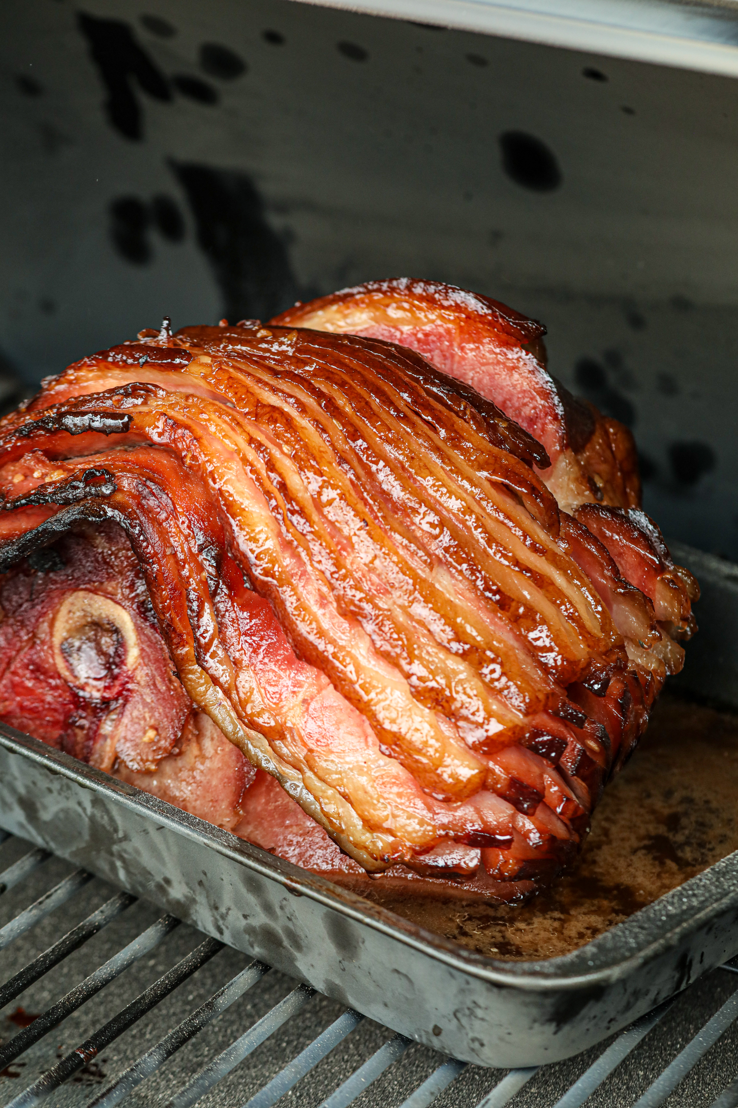 The ham has developed a beautifully caramelized crust, with the spiral-cut layers slightly crisped on the edges. The glaze has thickened and given the ham a glossy sheen, while the bone-in portion remains tender. The pan still holds some of the flavorful juices, ensuring the ham stays moist. The grill's heat has created a rich, smoky exterior, making this a visually stunning and deliciously prepared dish.