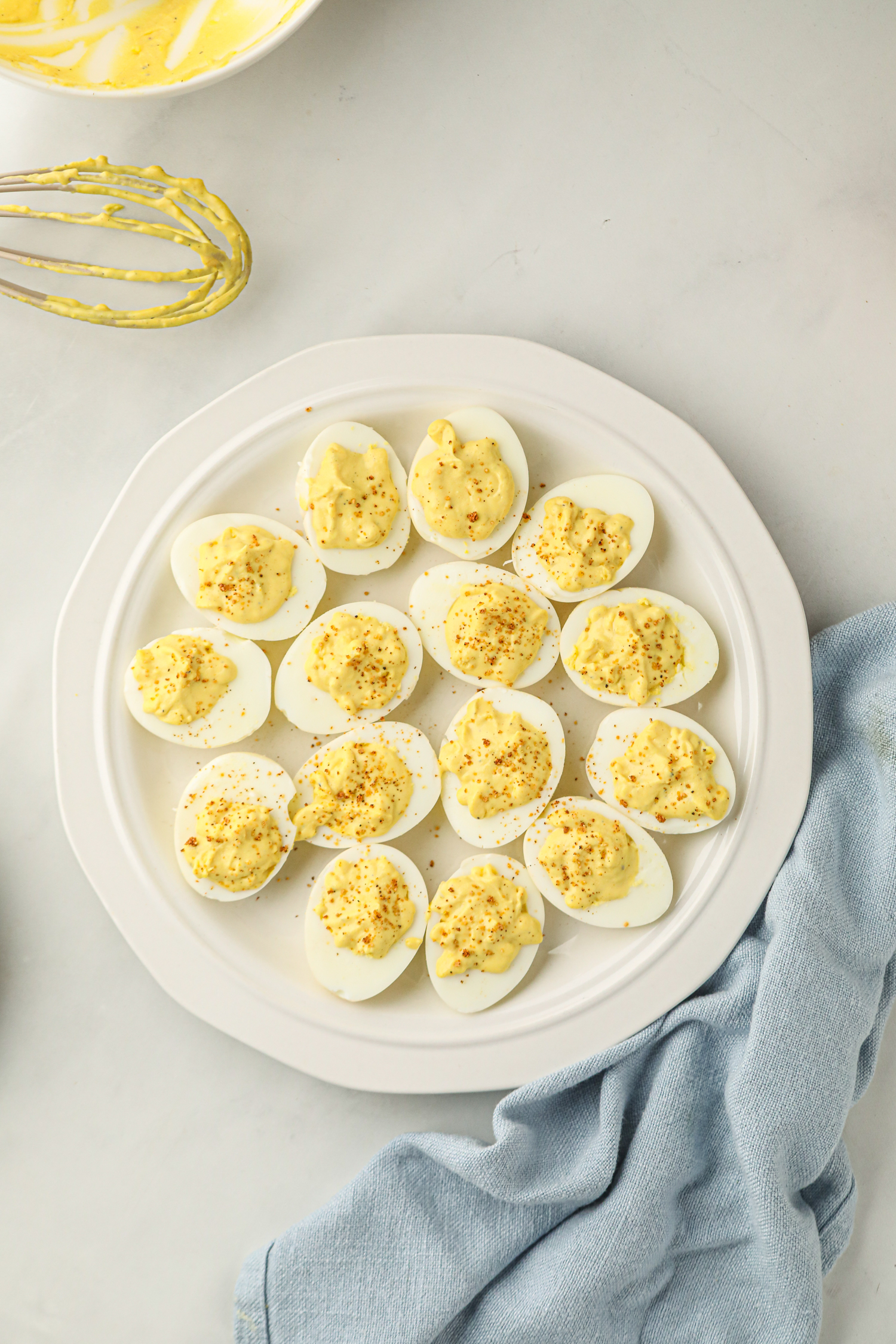 A white plate with smoked deviled eggs, each filled with a creamy yellow yolk mixture and sprinkled with seasoning. A whisk coated with the yolk mixture and a partially visible mixing bowl are in the background, along with a light blue cloth napkin. The dish appears freshly prepared and ready to be served.