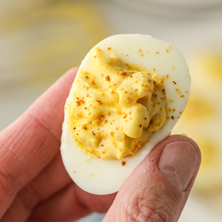 A close-up of a hand holding a smoked deviled egg. The egg white is filled with a creamy, yellow yolk mixture and sprinkled with seasoning. The texture of the filling appears smooth with a slight rustic consistency. The background is blurred, showing more deviled eggs on a plate.