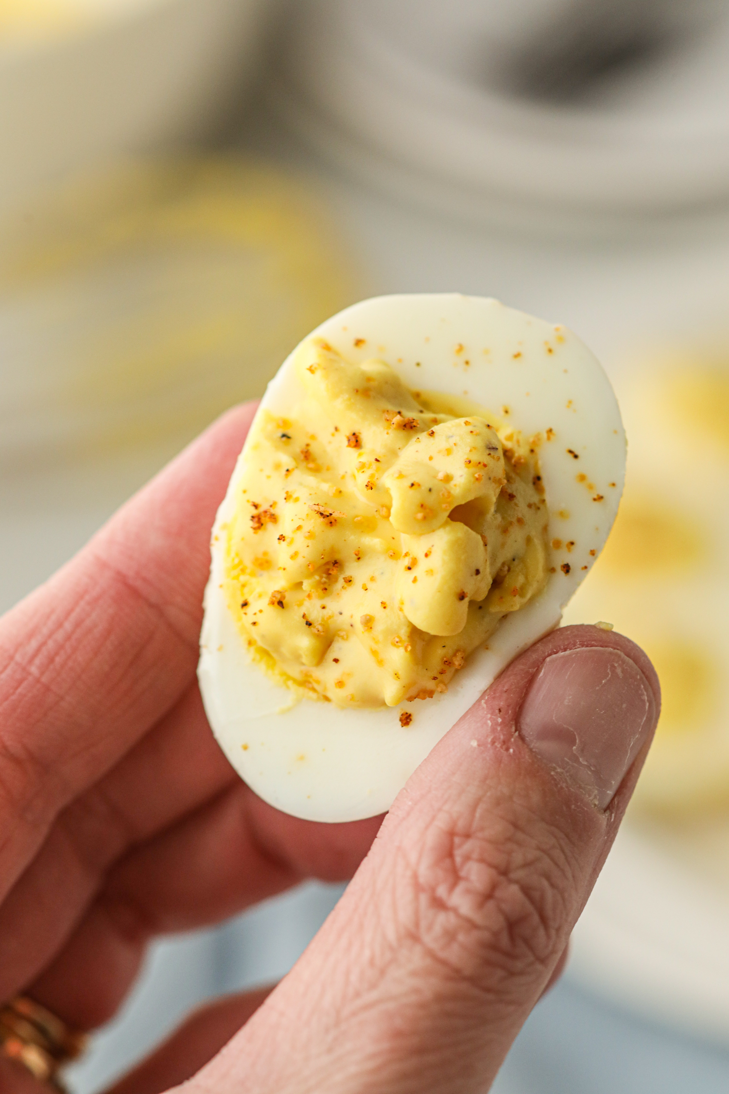 A close-up of a hand holding a smoked deviled egg. The egg white is filled with a creamy, yellow yolk mixture and sprinkled with seasoning. The texture of the filling appears smooth with a slight rustic consistency. The background is blurred, showing more deviled eggs on a plate.