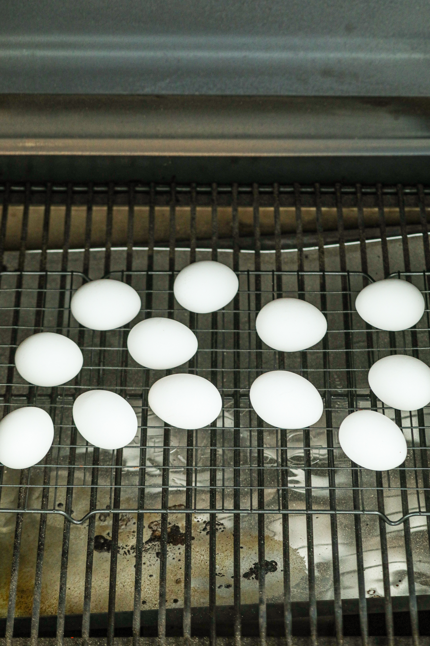 The image shows several white eggs placed on a wire rack inside a smoker or grill. The eggs are arranged in a scattered manner, and the grill appears to have some charred residue on the bottom. The setup suggests that the eggs are being smoked, likely for a recipe such as smoked deviled eggs.