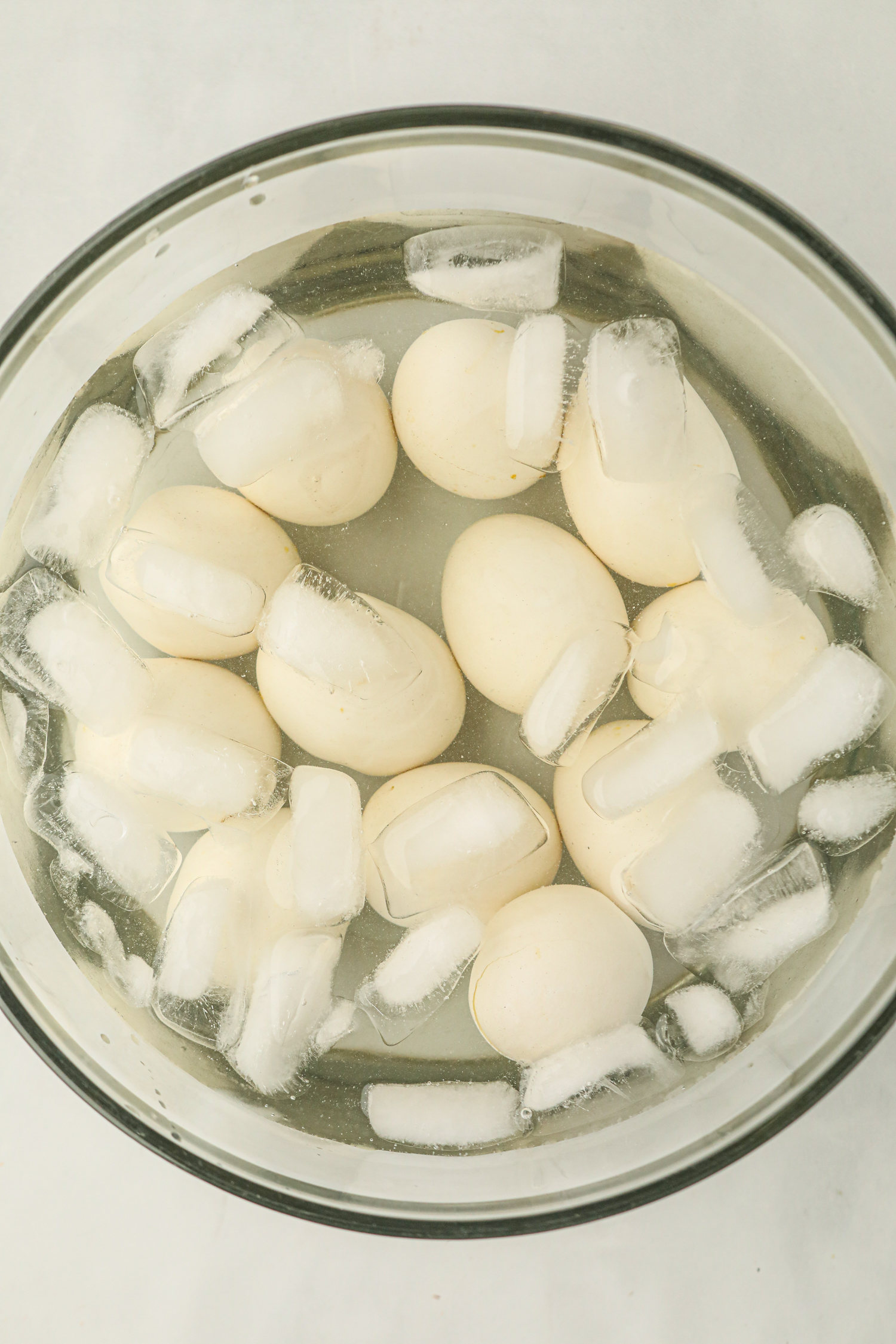 A glass bowl filled with smoked eggs in an ice water bath. The eggs have a slightly off-white or lightly tanned appearance, indicating they have been smoked. Ice cubes float around the eggs, helping to cool them quickly, likely to stop the cooking process and make peeling easier.