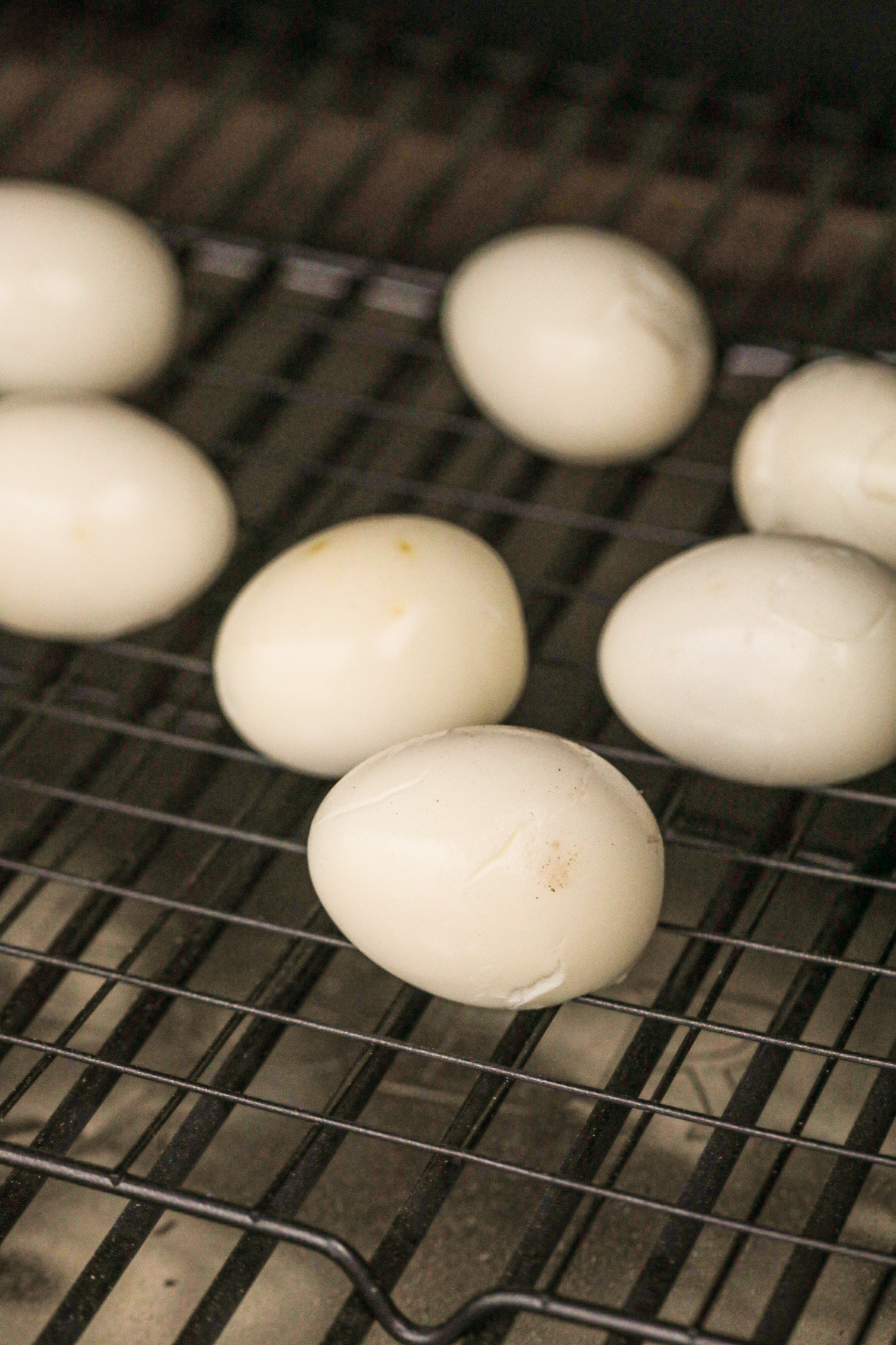 Peeled hard-boiled eggs placed on a wire rack inside a smoker. The eggs have a slightly golden or off-white hue, indicating they have been smoked. Some eggs show minor surface cracks and texture changes from the smoking process.