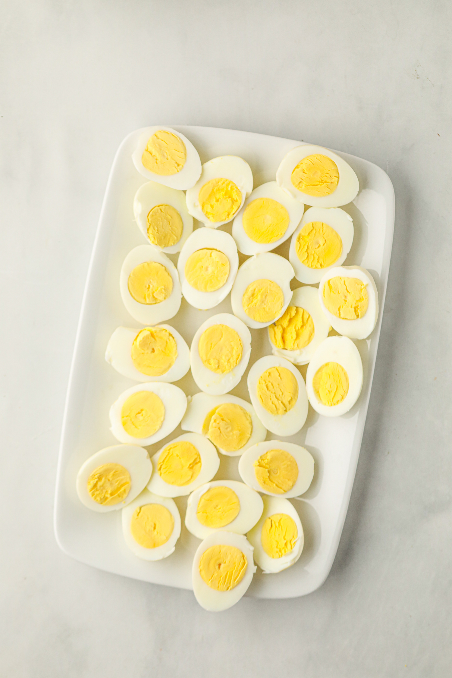 A white rectangular plate filled with smoked hard-boiled eggs that have been cut in half. The eggs have a slightly firm, bright yellow yolk and smooth white exterior, arranged neatly on the plate. They are likely prepared for making smoked deviled eggs.