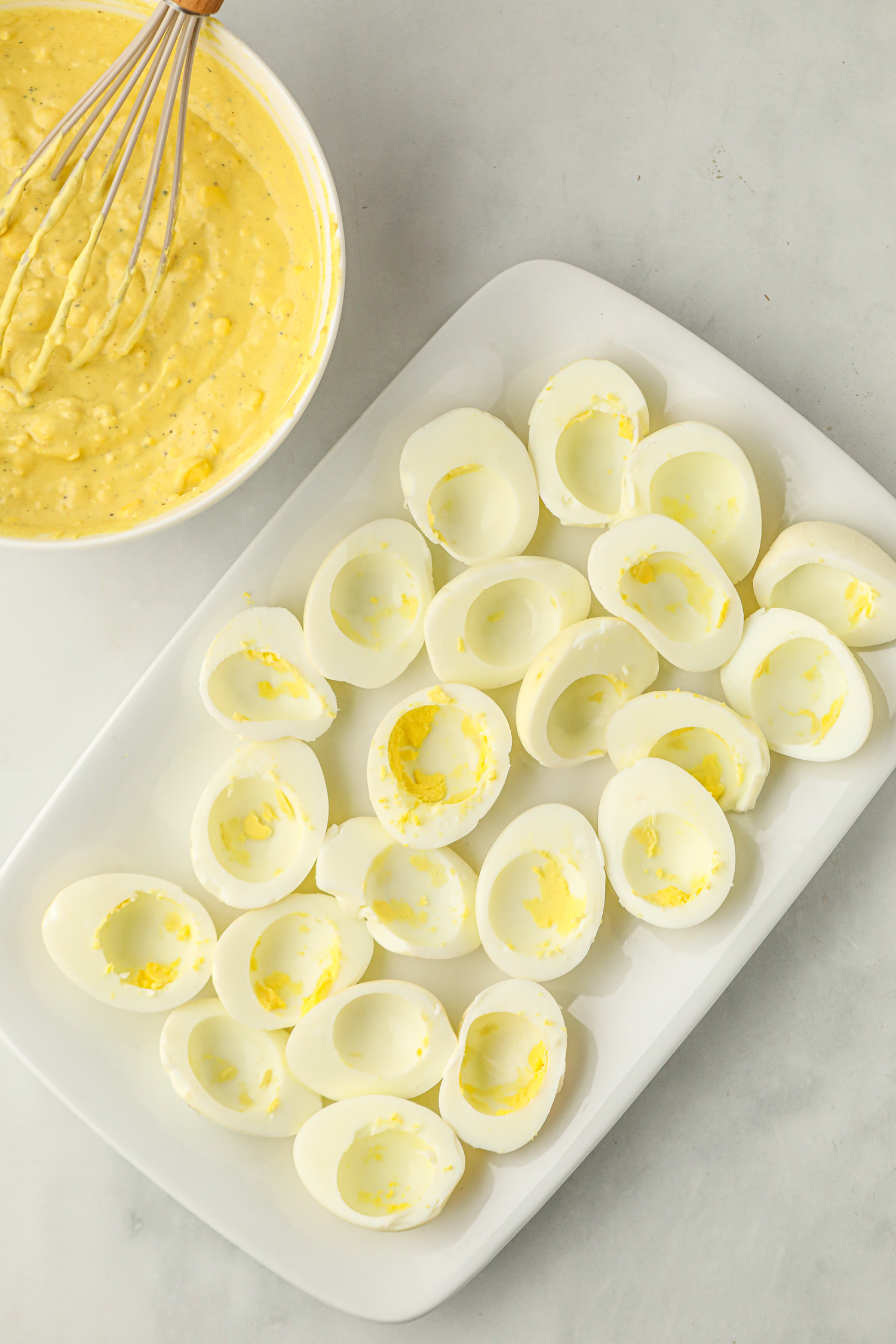 A white rectangular plate filled with halved smoked eggs with their yolks removed, leaving hollow centers. Beside the plate, a bowl contains a creamy, yellow deviled egg filling, with a whisk partially submerged. The image suggests the preparation stage before stuffing the egg whites with the yolk mixture.