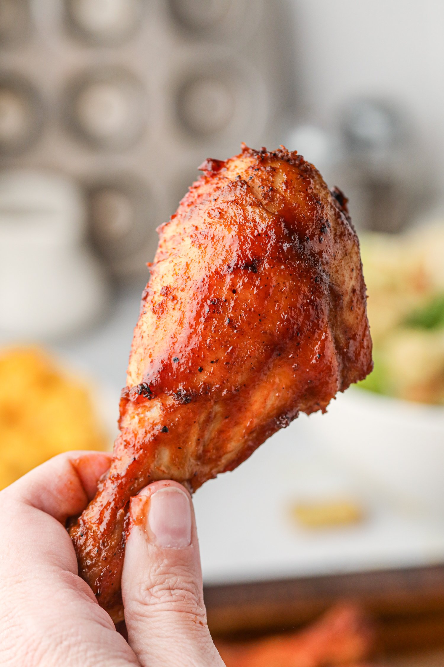 A hand holding a single glazed BBQ chicken drumstick, close up, showing the crispy, caramelized skin with visible seasoning and sauce.

