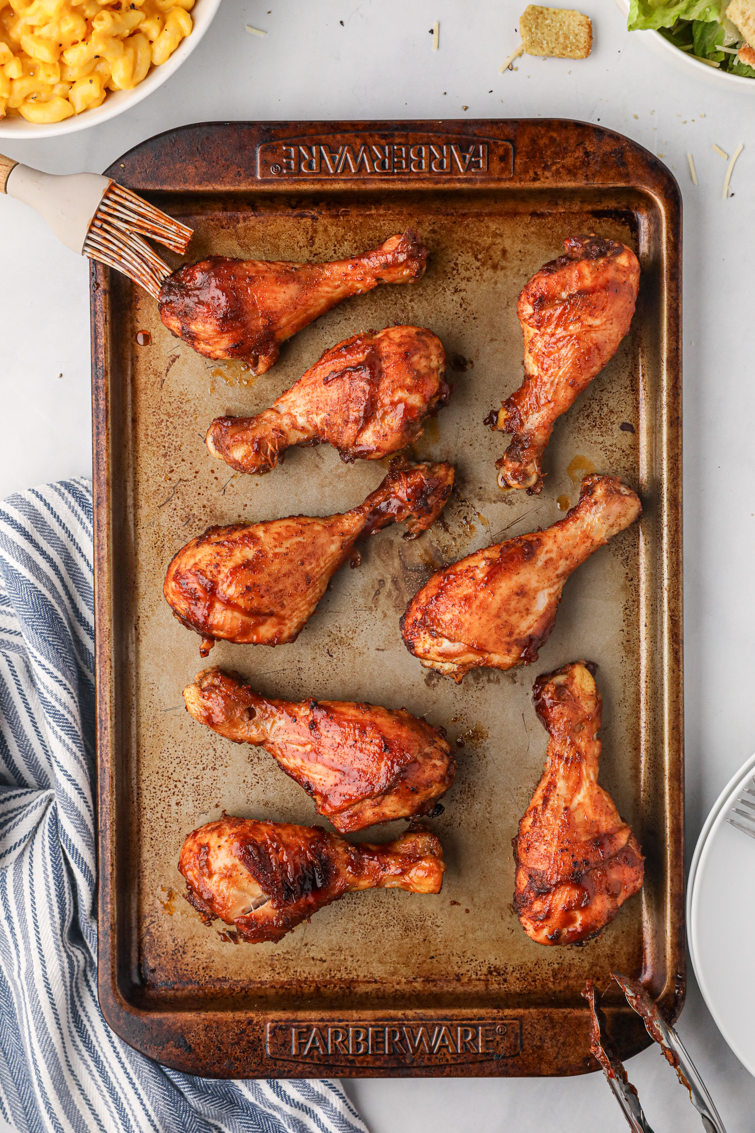 A baking sheet filled with eight cooked BBQ chicken drumsticks, some with grill marks and a glossy finish, sitting on a countertop near a striped kitchen towel and side dishes including macaroni and cheese and salad.