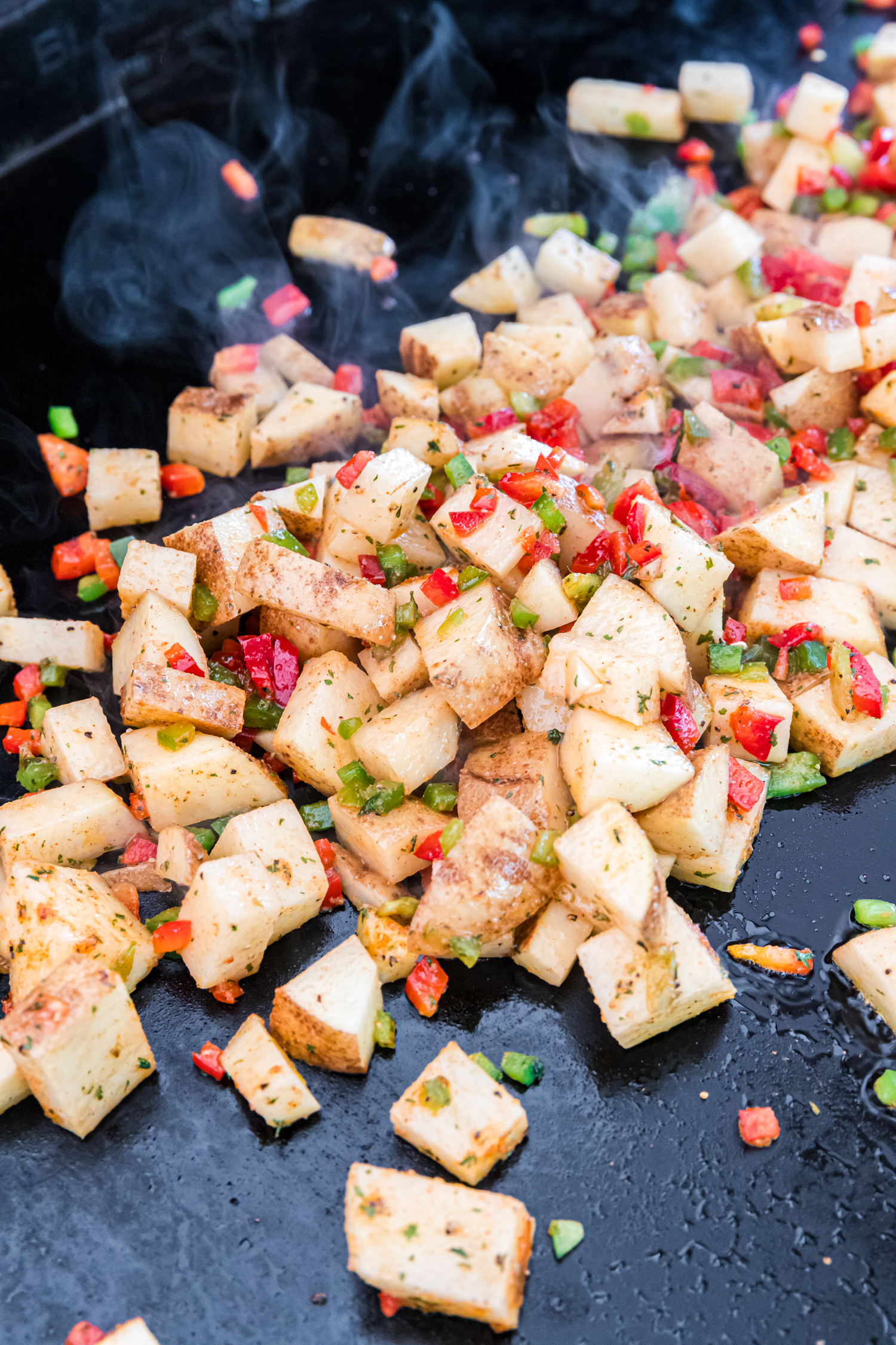 Seasoned potatoes and diced peppers sizzling on a hot Blackstone griddle with visible steam.