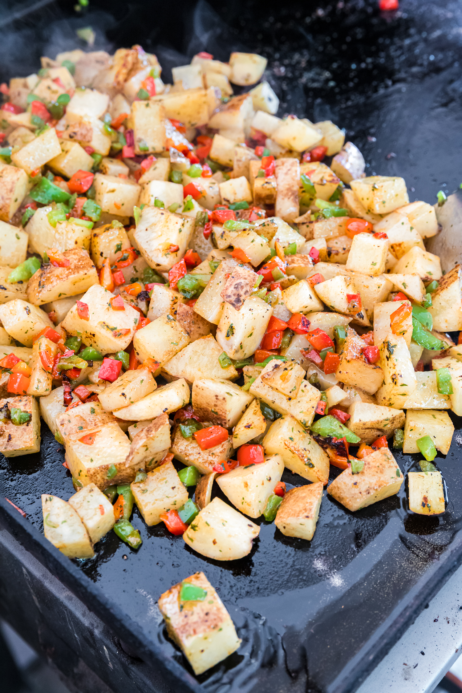 Golden brown breakfast potatoes cooking on a flat-top griddle with red and green bell peppers.
