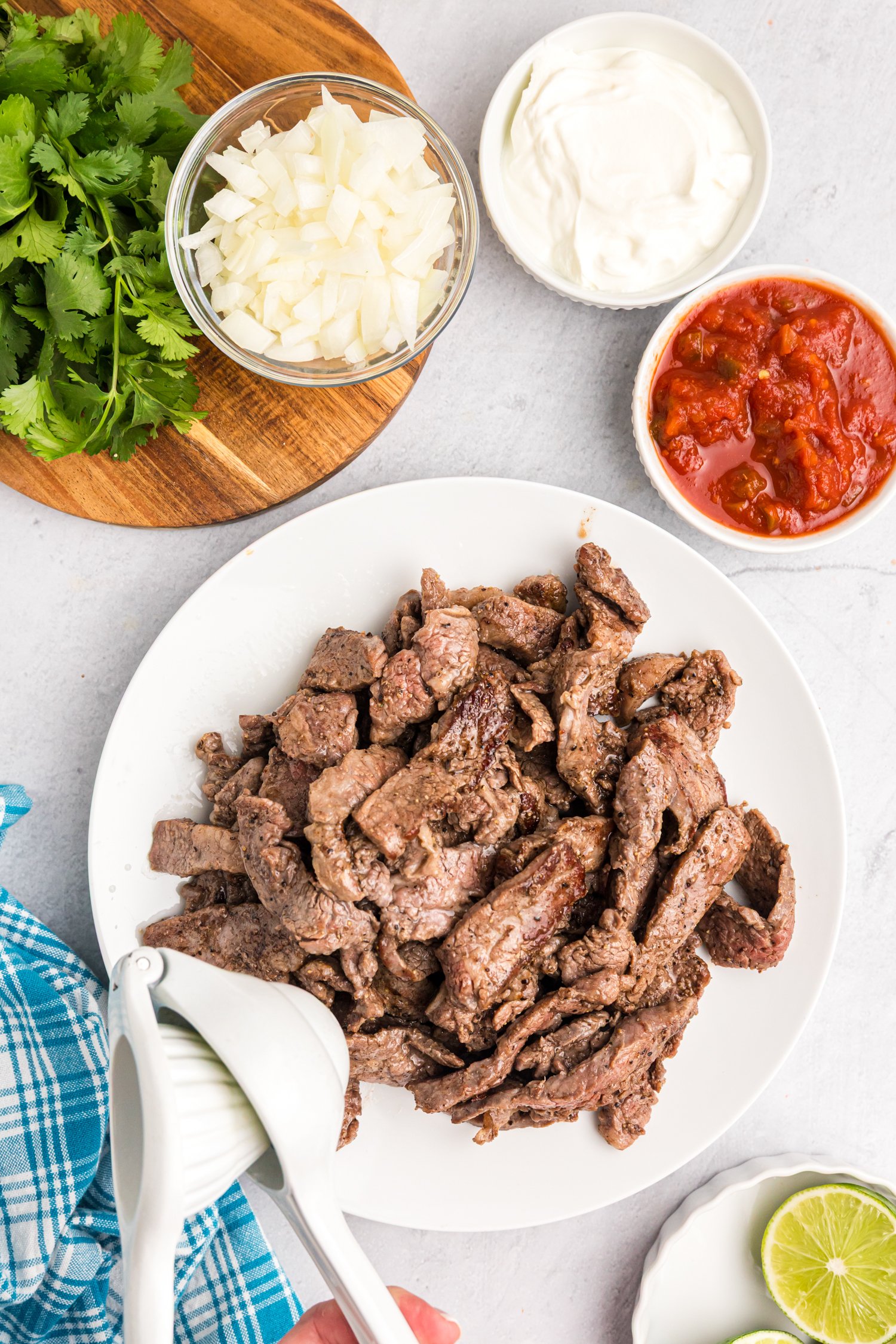 A plate of cooked beef strips with a hand squeezing fresh lime juice over them, accompanied by bowls of chopped onions, cilantro, salsa, and sour cream.