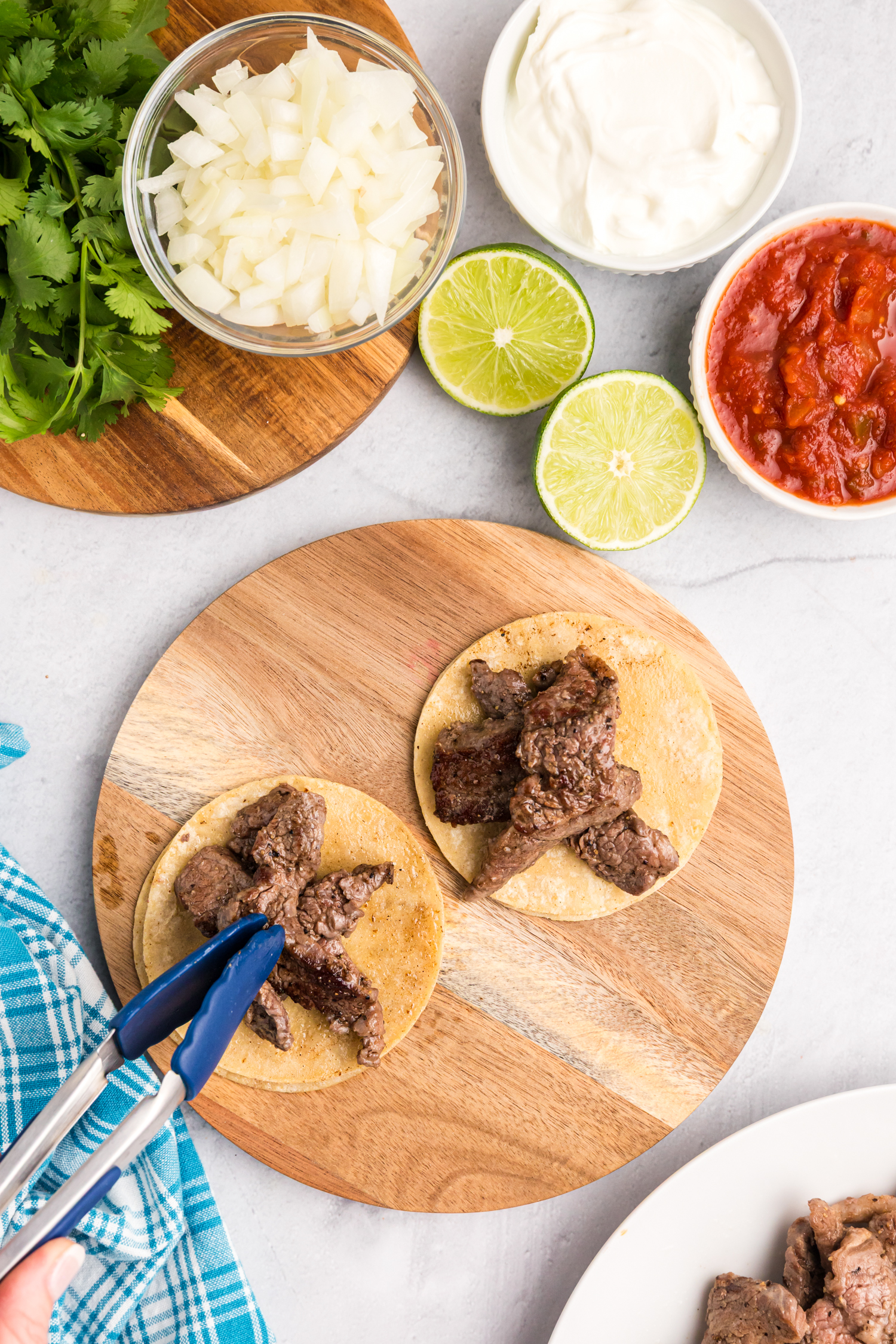 A wooden board with two small street tacos being assembled, each topped with cooked beef, with bowls of toppings in the background.