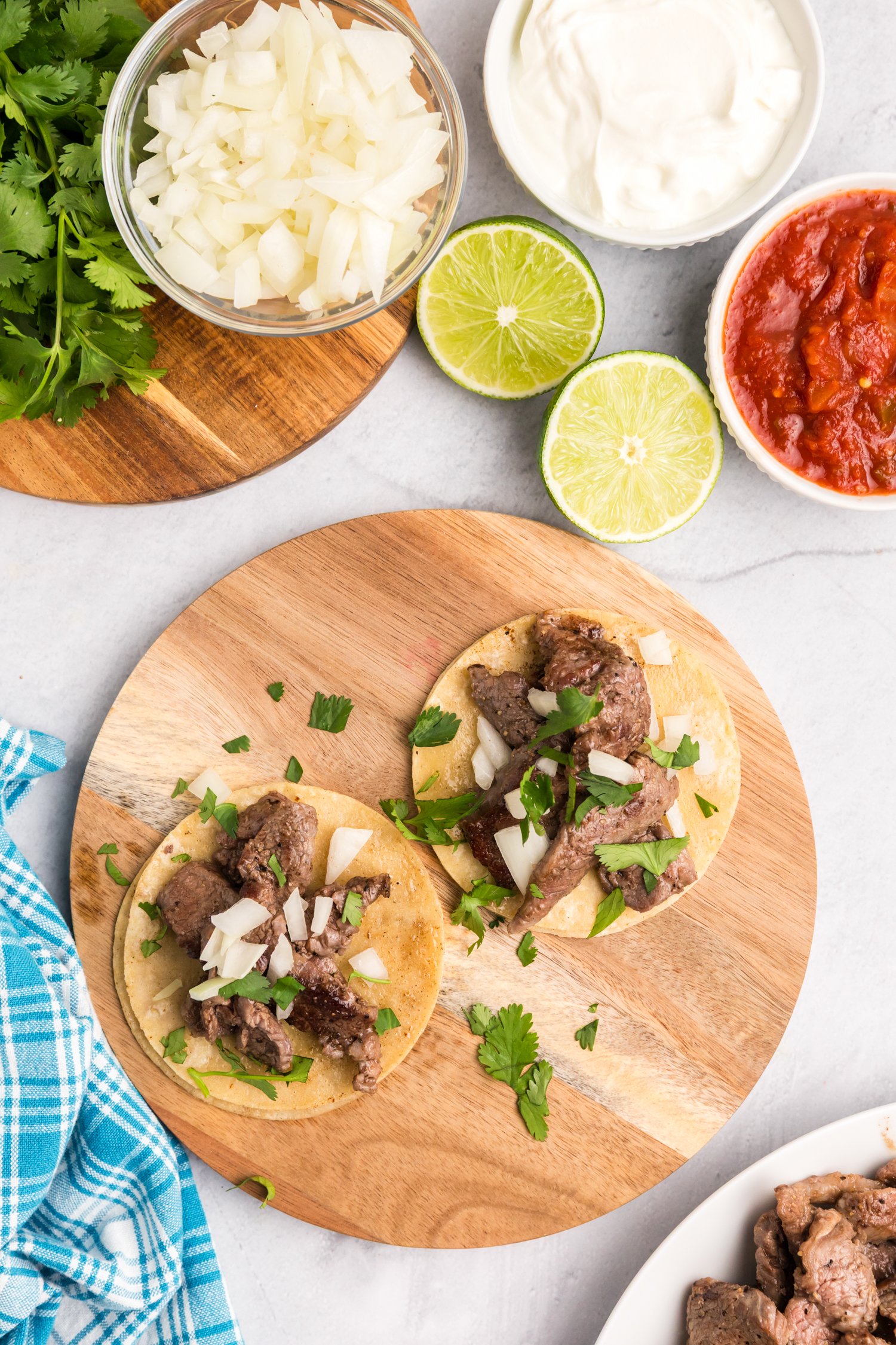 Two small street tacos on a wooden board, topped with seasoned beef, diced onions, and fresh cilantro, with lime halves, sour cream, and salsa nearby.