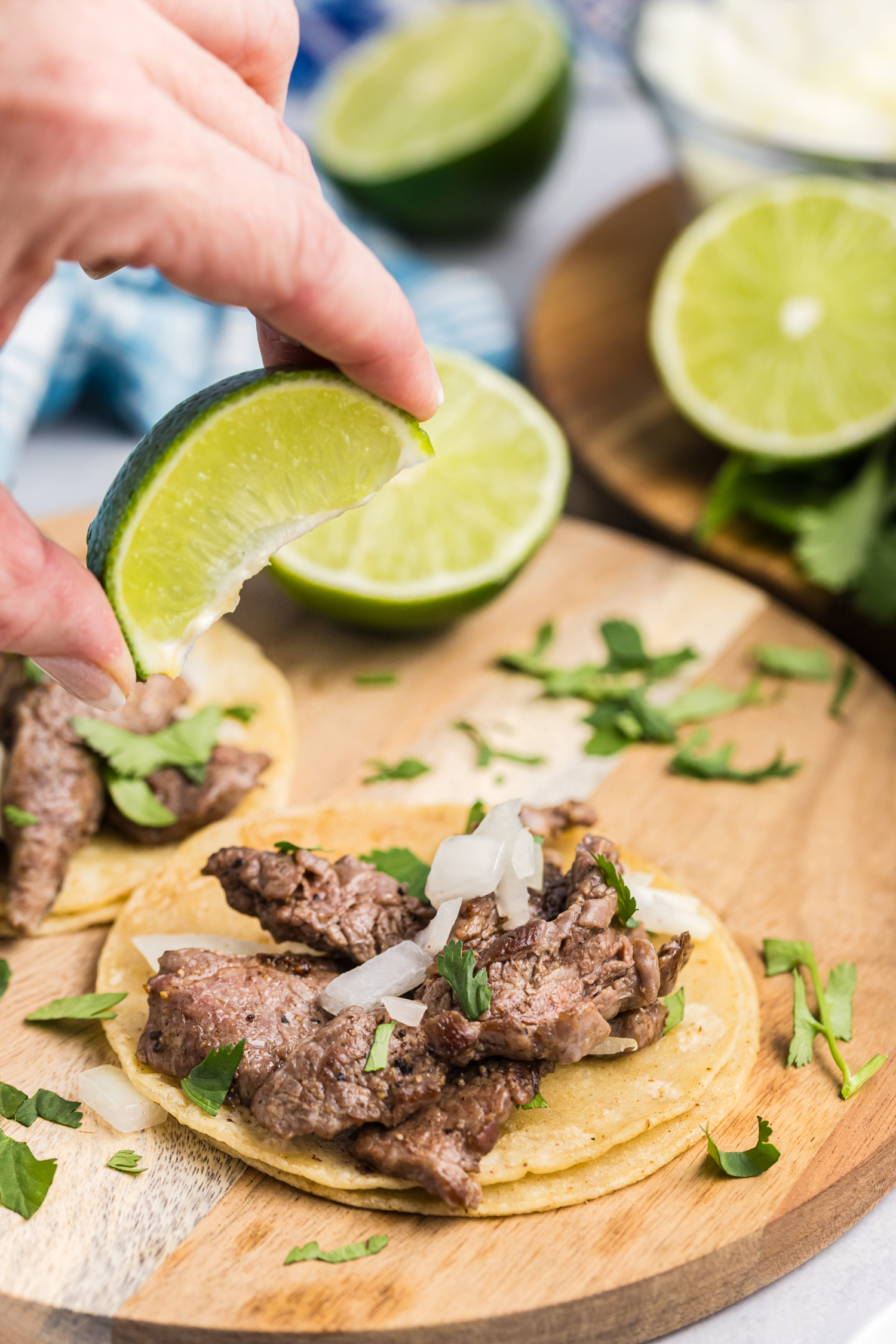A hand squeezing a fresh lime wedge over two beef street tacos, garnished with onions and cilantro on a wooden board.