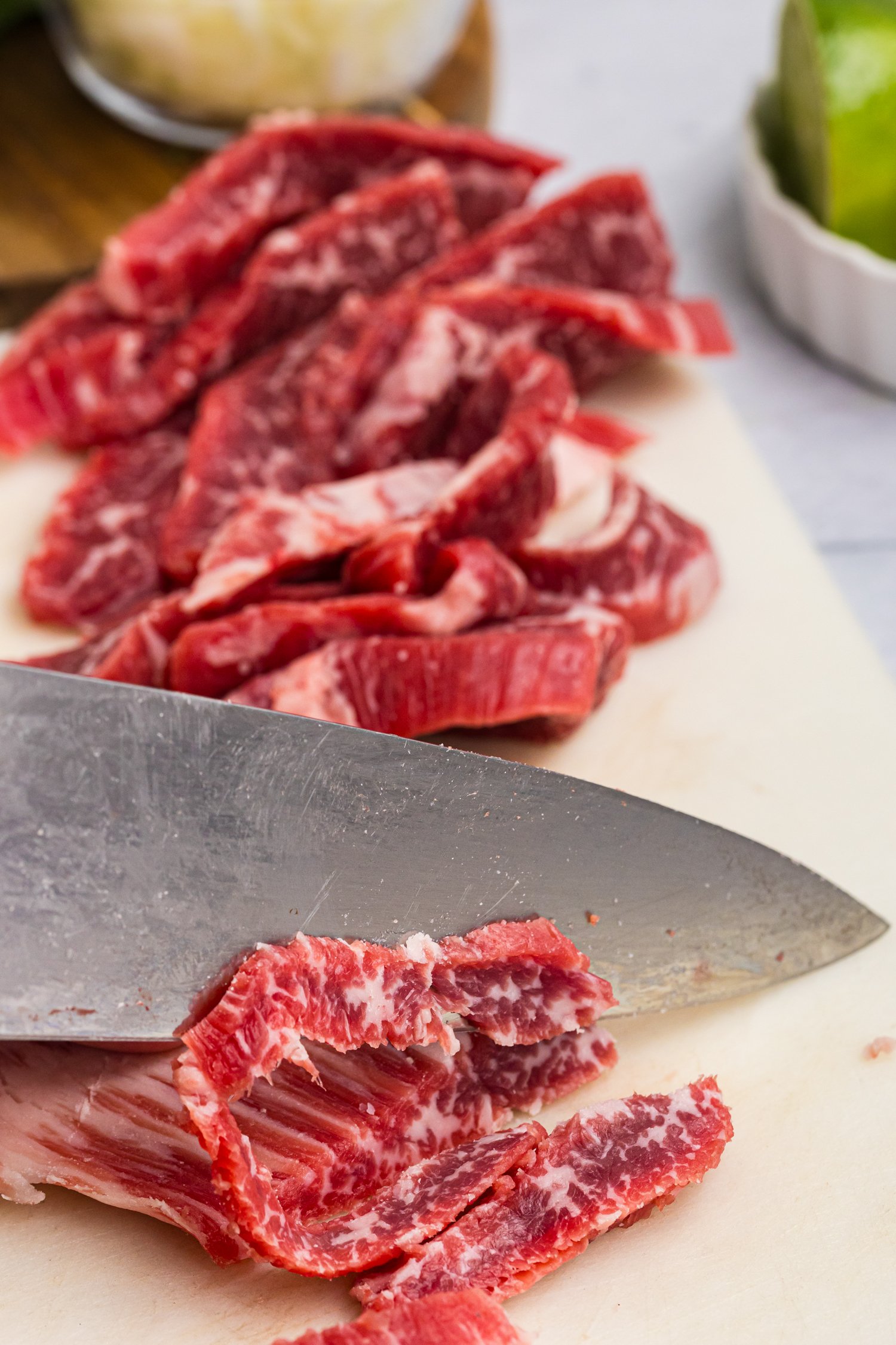 A close-up of a sharp knife slicing through marbled raw beef strips on a cutting board.