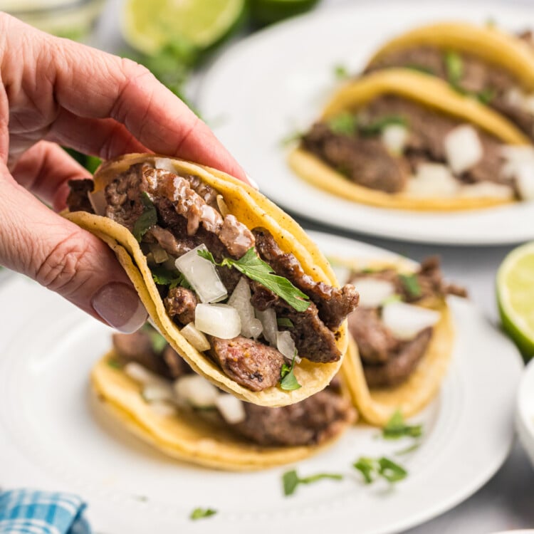 A hand holding a fully assembled street taco filled with seasoned beef, onions, and cilantro, with more tacos and toppings in the background.