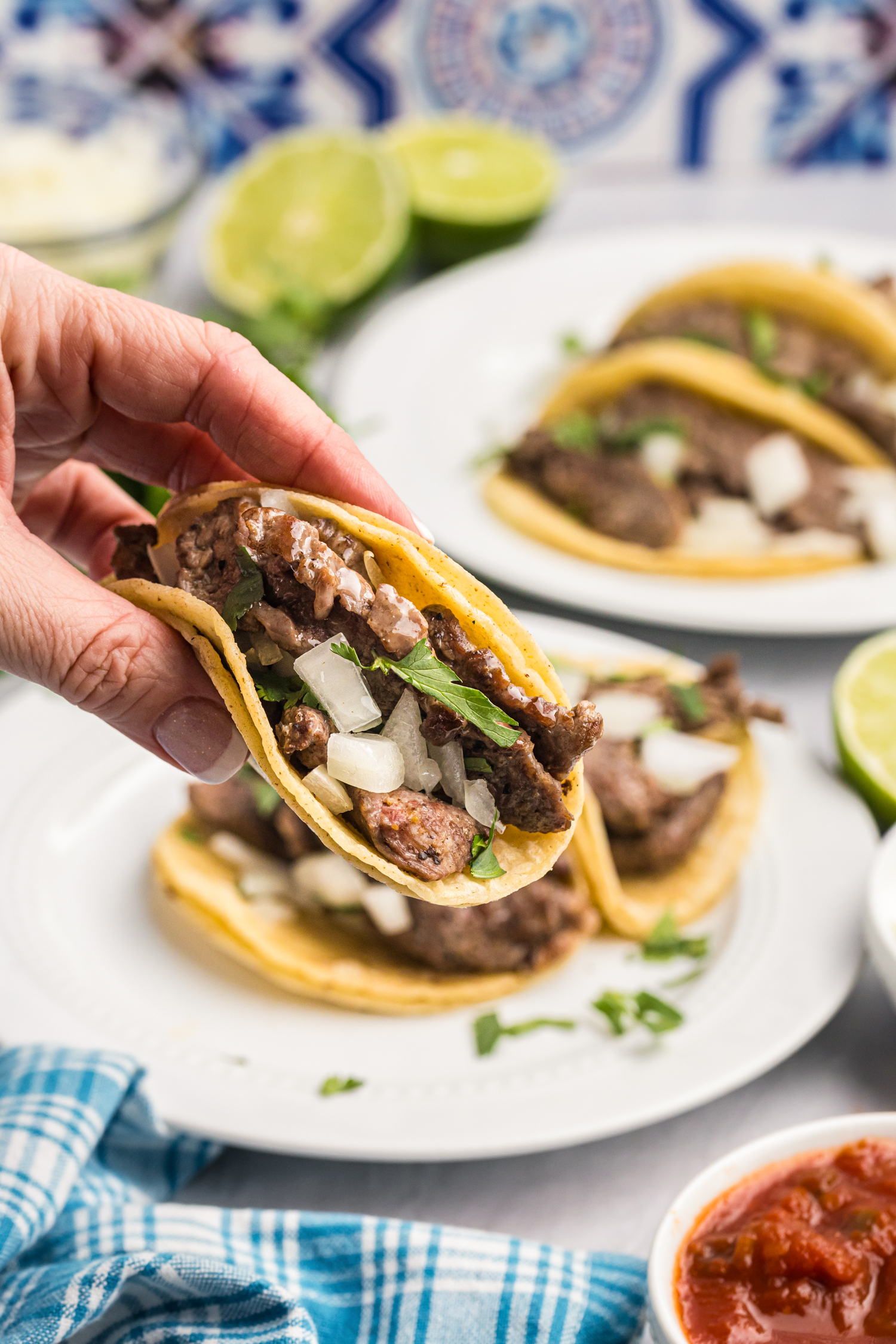 A hand holding a fully assembled street taco filled with seasoned beef, onions, and cilantro, with more tacos and toppings in the background.