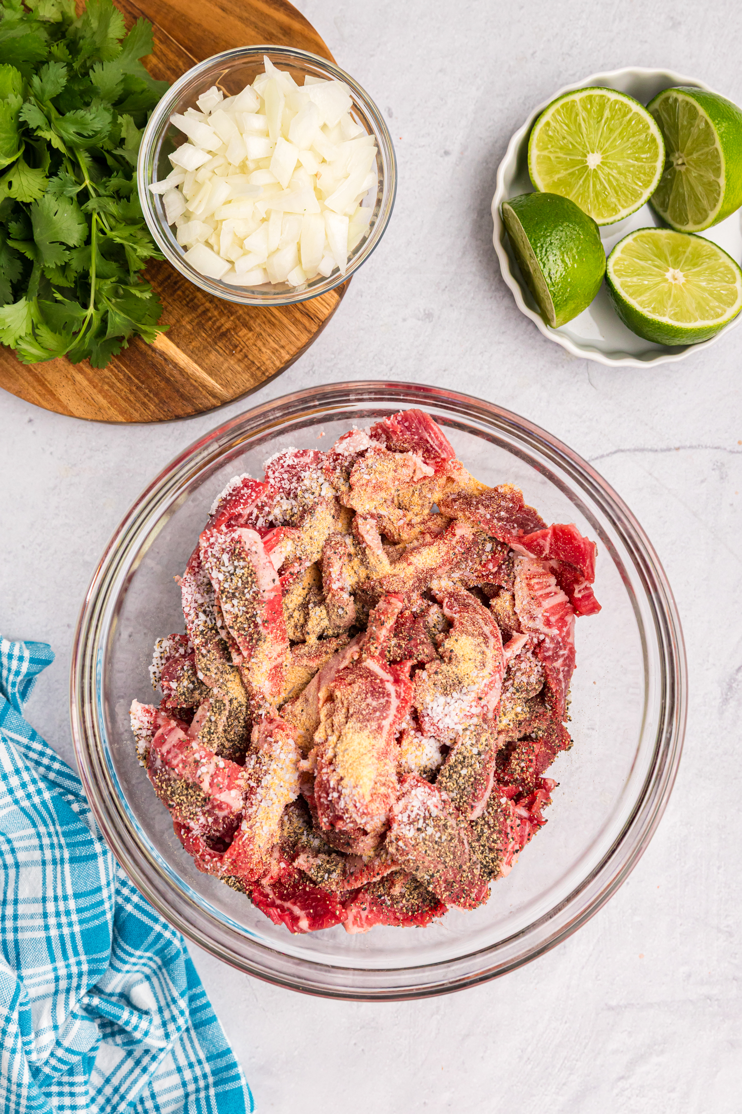 A glass bowl filled with raw beef strips covered in seasoning, surrounded by chopped onions, cilantro, and fresh limes.