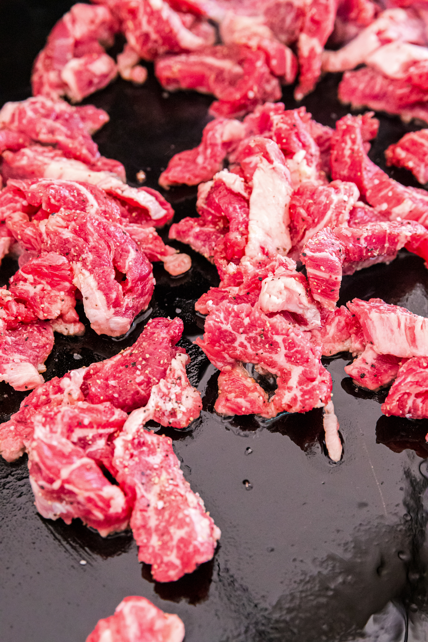 Raw, seasoned beef strips sizzling on a hot griddle with a slight sear forming.