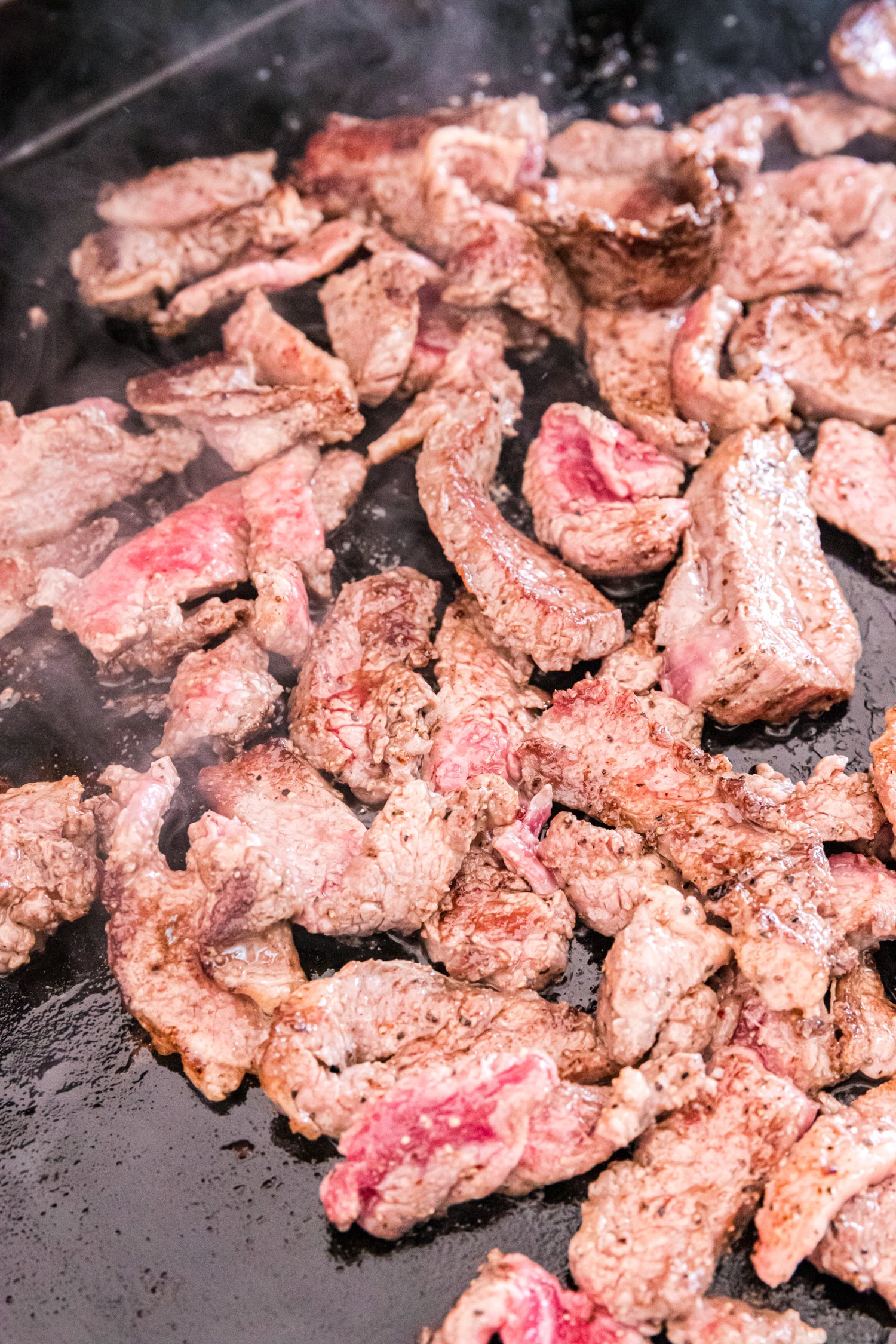 Beef strips beginning to brown and cook through on a griddle, releasing steam.