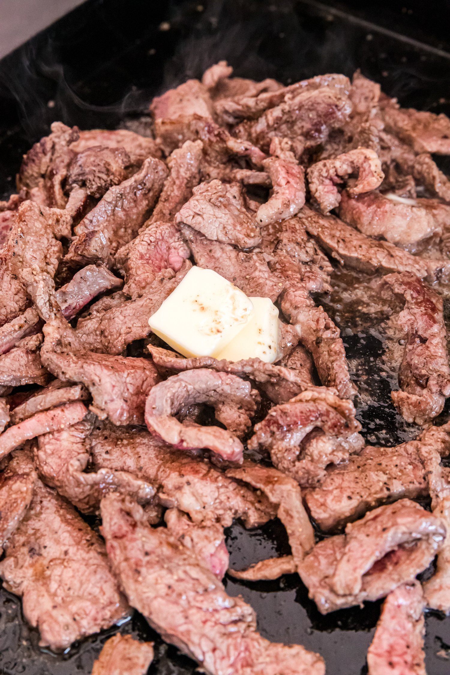 Fully cooked, browned beef strips on the griddle with a pat of butter melting on top.