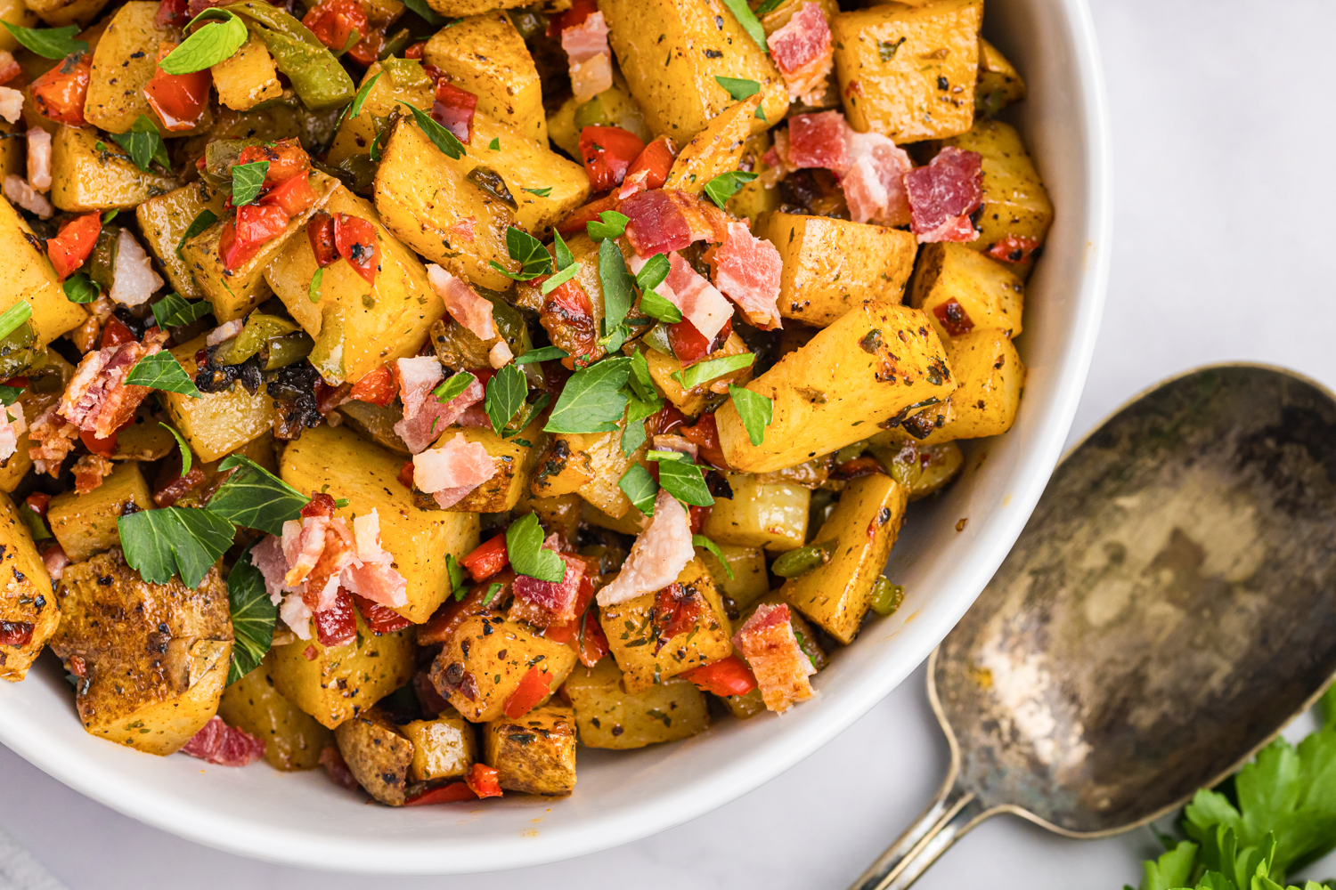 Close-up of crispy Blackstone breakfast potatoes with bacon, red and green bell peppers, and fresh parsley in a white serving bowl.