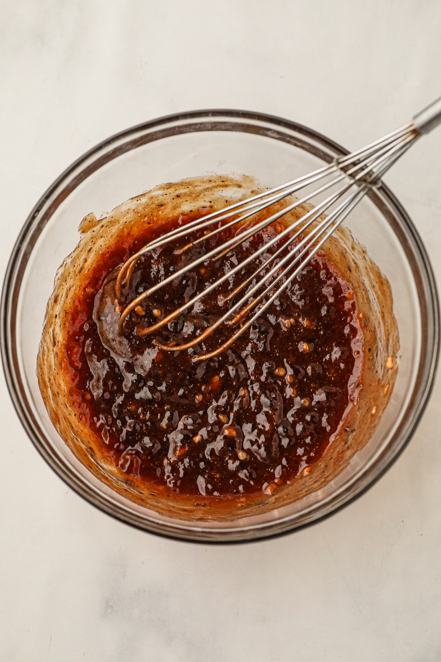 A bowl of dark reddish BBQ sauce mixture being whisked, showing visible pepper and garlic pieces in the thick glaze.