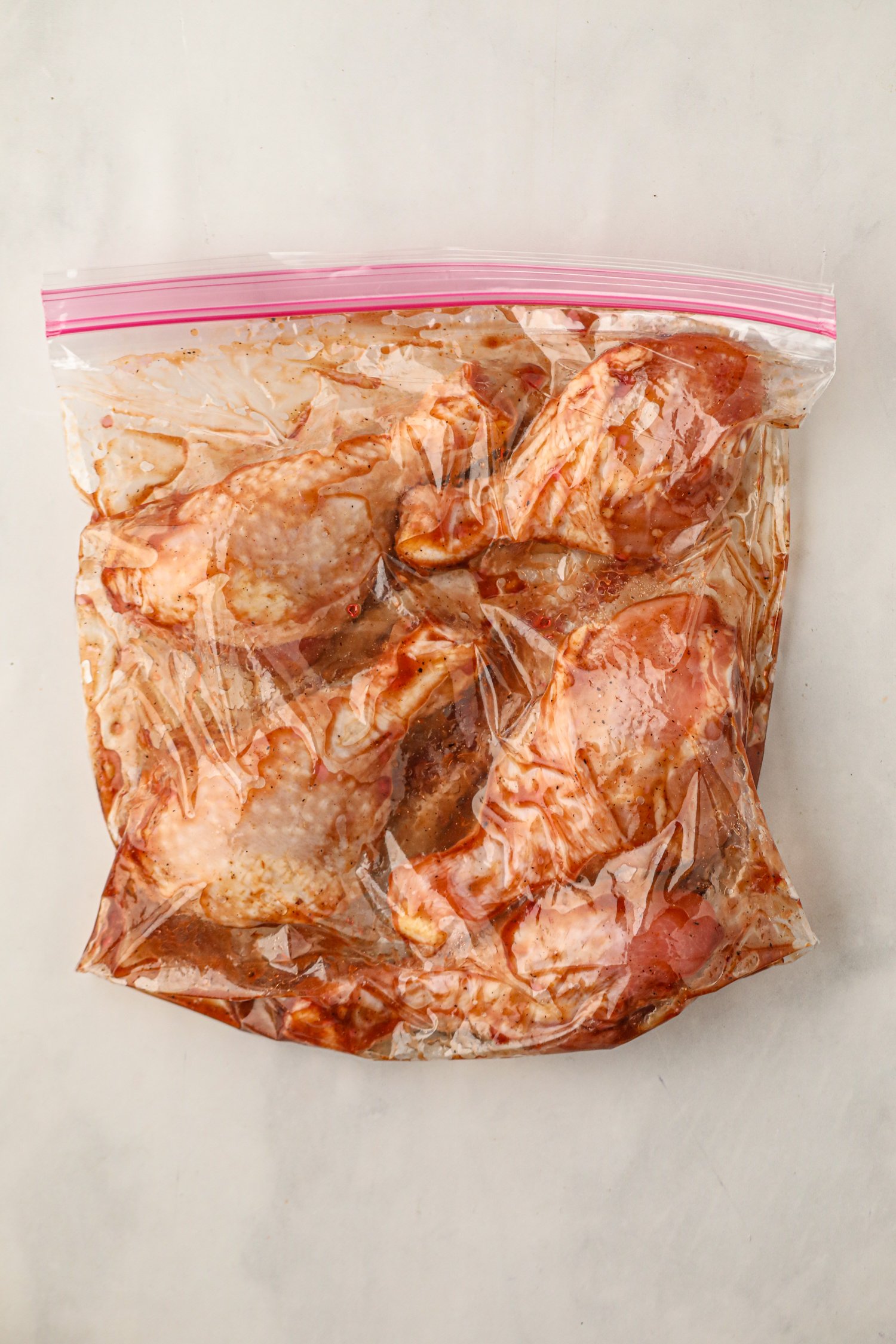 A sealed plastic ziplock bag filled with raw chicken drumsticks marinating in a reddish BBQ sauce, laid flat on a white surface.