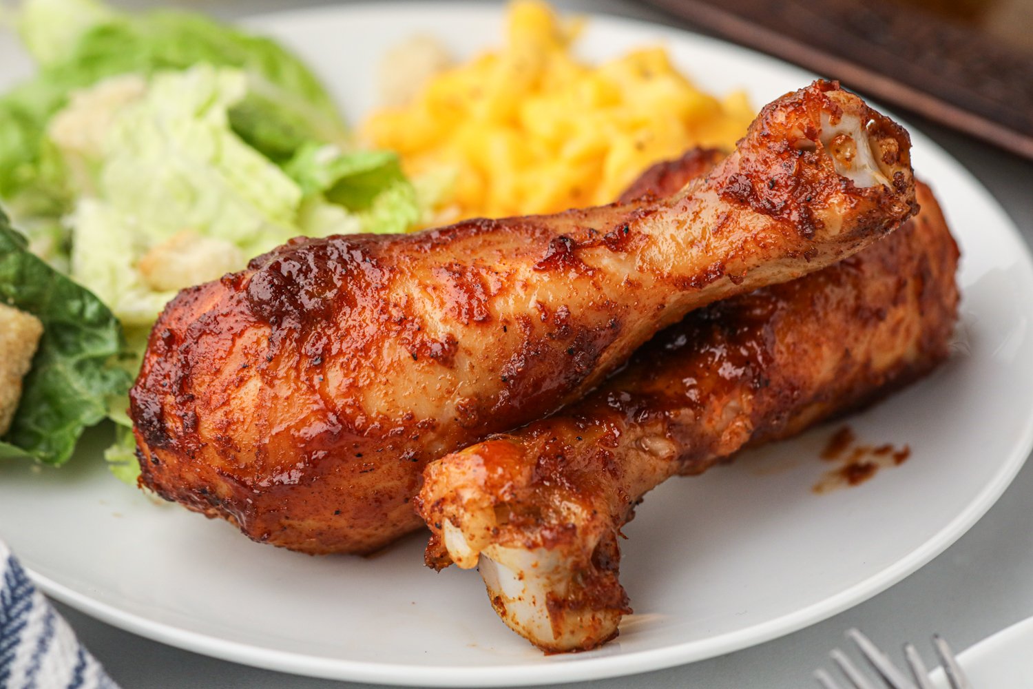 A plate of two glazed BBQ chicken drumsticks served with a side of macaroni and cheese and Caesar salad, close-up view showing juicy and saucy chicken skin.