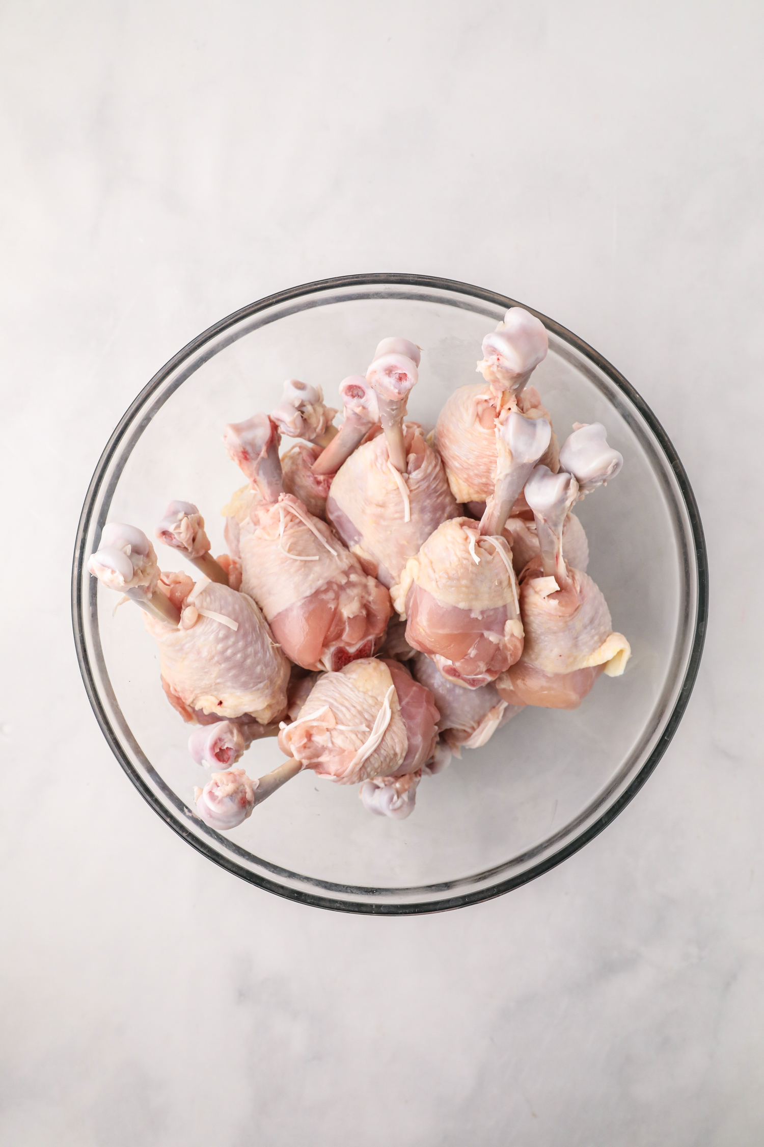 Raw chicken drumsticks placed upright in a clear glass bowl on a light countertop.