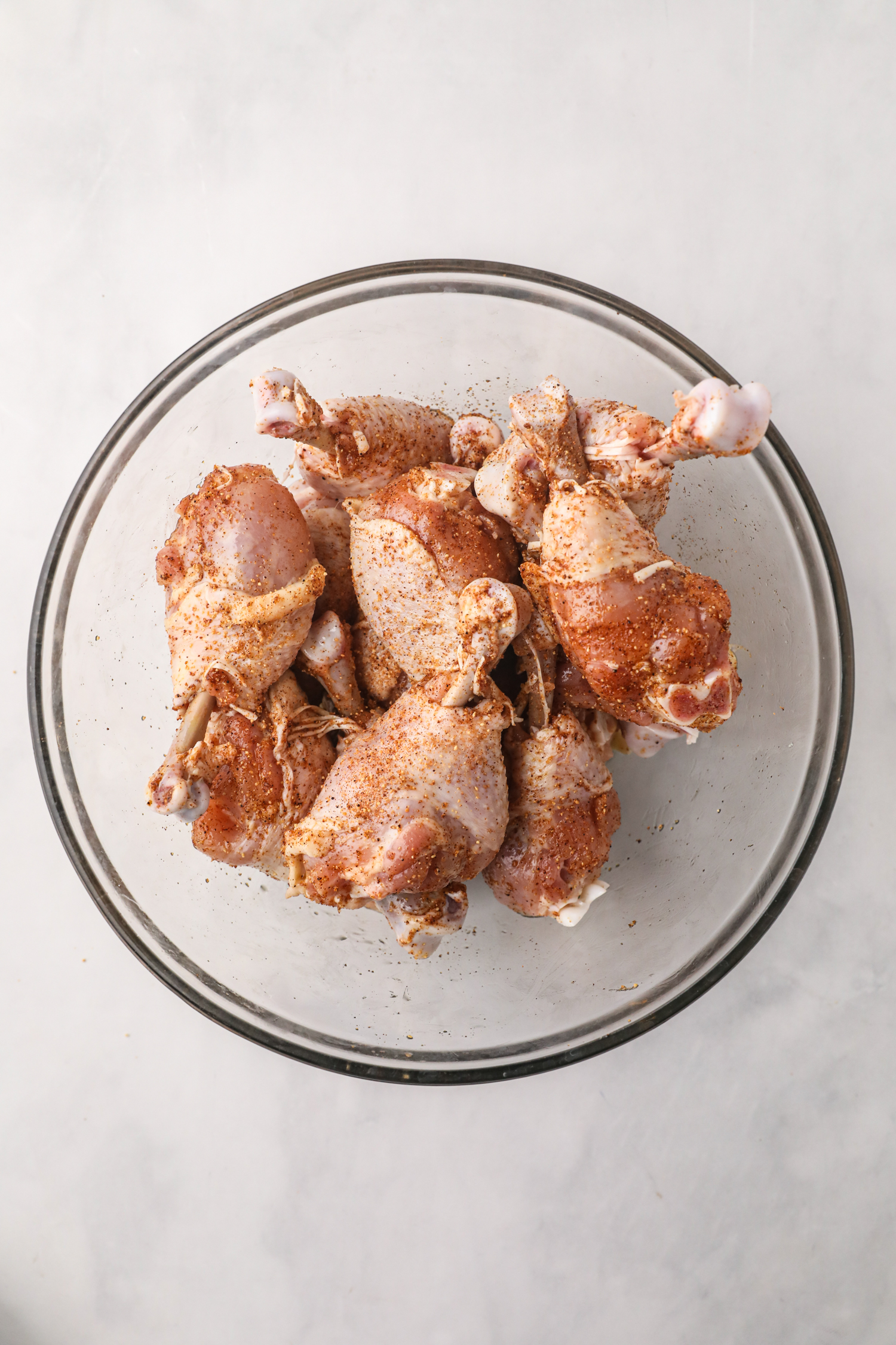 Chicken drumsticks in a glass bowl, evenly coated with BBQ rub seasoning.
