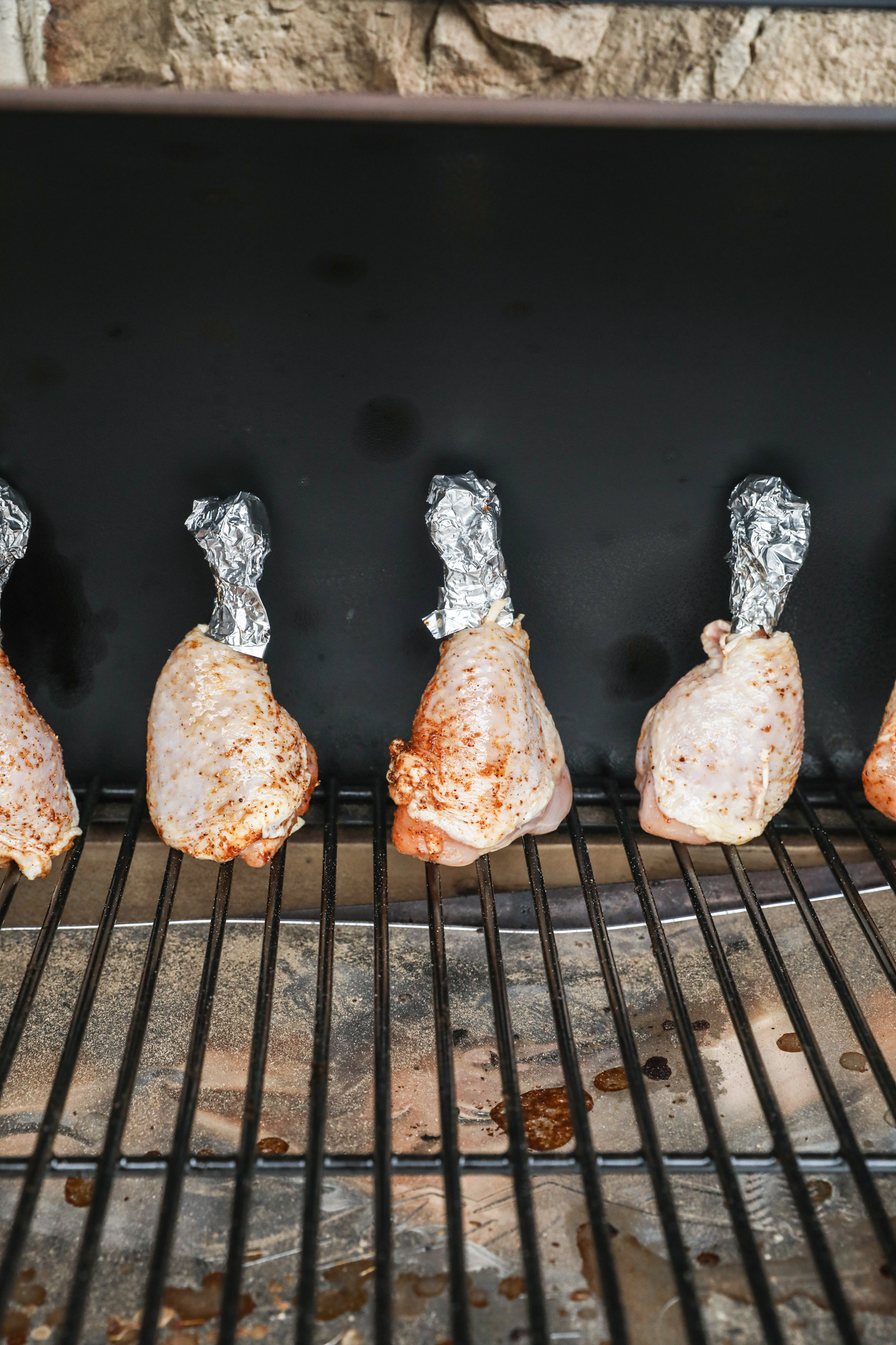 Chicken drumsticks standing upright inside a smoker, with foil-wrapped bone ends facing up.