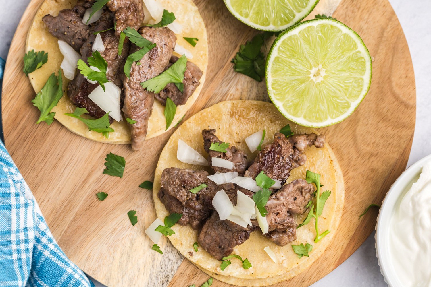 A top-down view of two street tacos on a wooden board, topped with beef, onions, and cilantro, with two halved limes and a bowl of sour cream nearby.