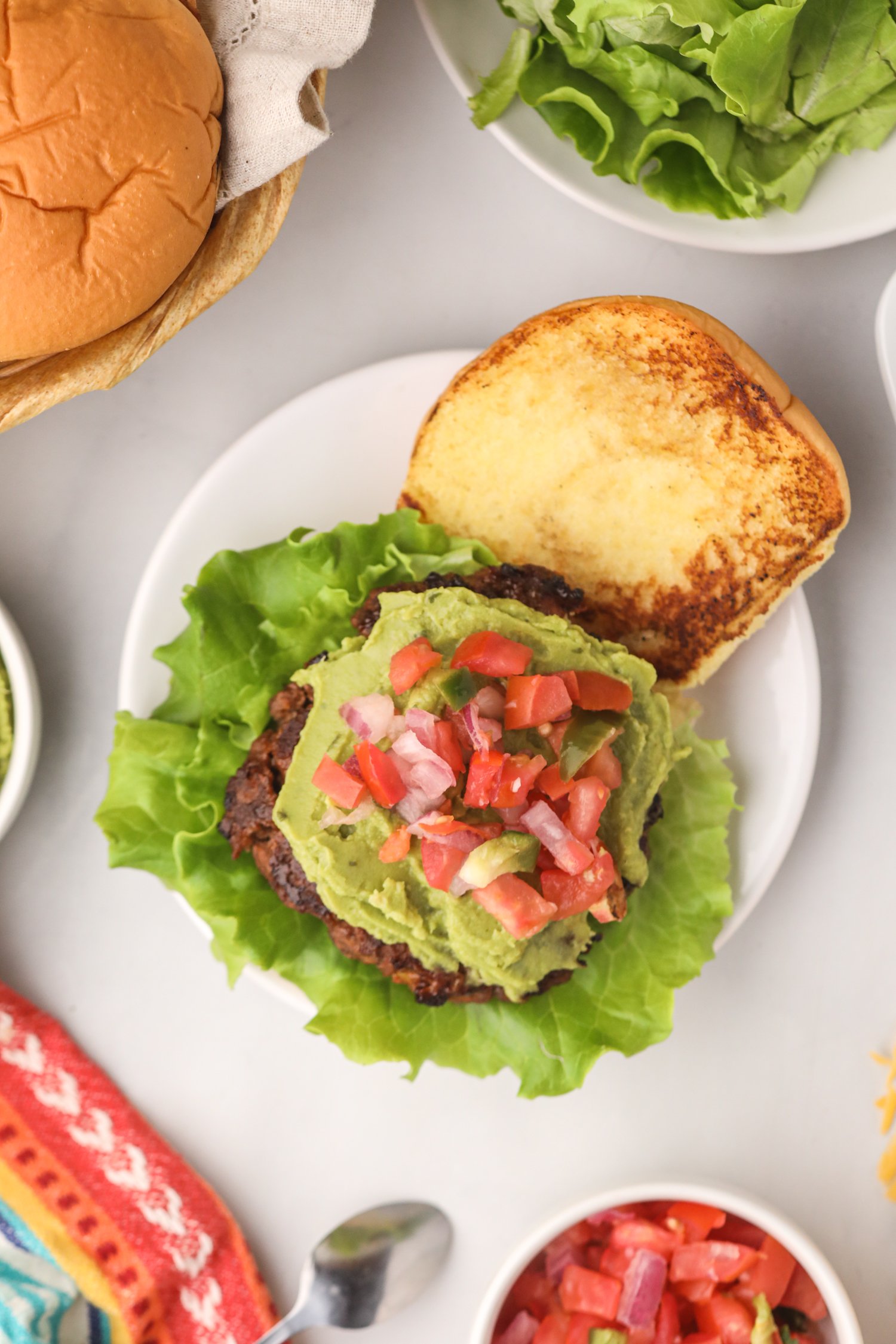 Open-faced taco burger on a plate with a grilled bun, fresh lettuce, guacamole, and pico de gallo.