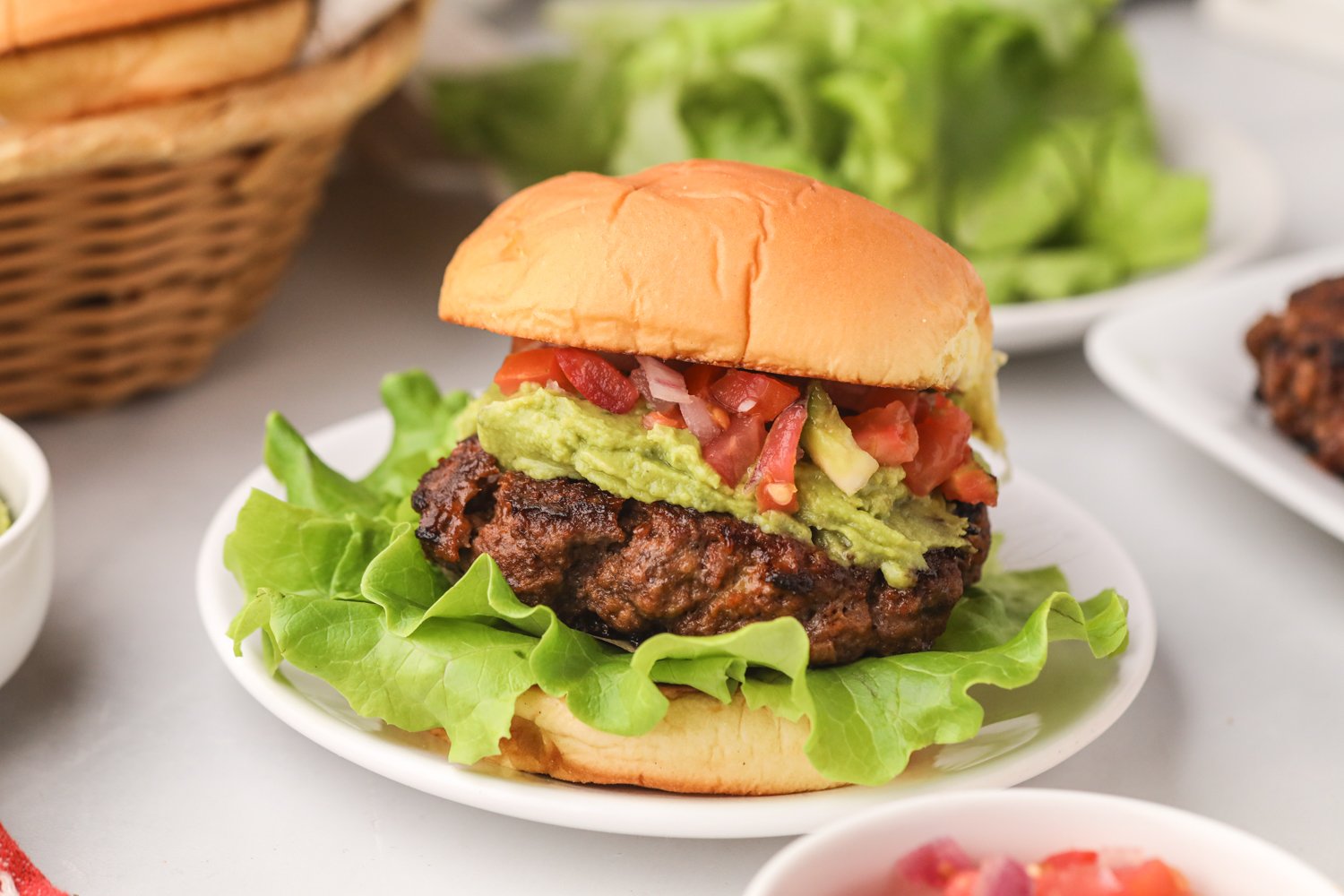 Fully assembled taco burger on a plate with a soft bun, leafy lettuce, guacamole, and a colorful mix of diced tomatoes, onions, and peppers.