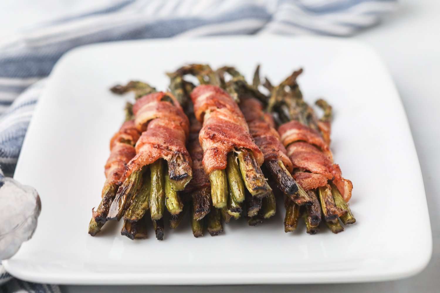A plate of grilled bacon-wrapped asparagus bundles, perfectly crisp and charred, arranged neatly on a white square plate with a striped napkin in the background.