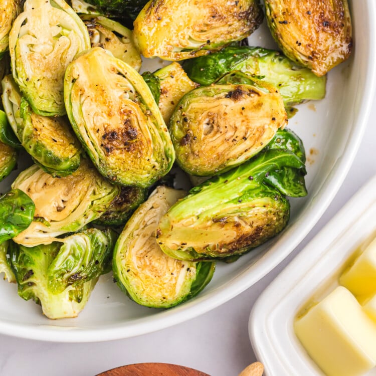 Golden-brown Brussels sprouts in a serving bowl with a dish of butter and brown sugar on the side.