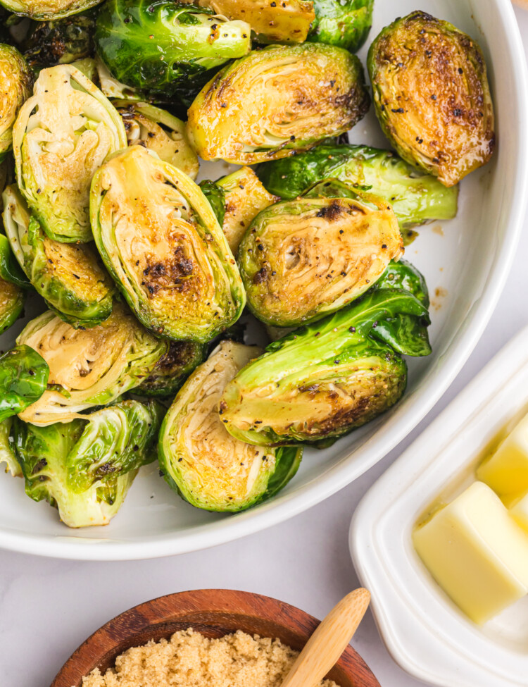 Golden-brown Brussels sprouts in a serving bowl with a dish of butter and brown sugar on the side.