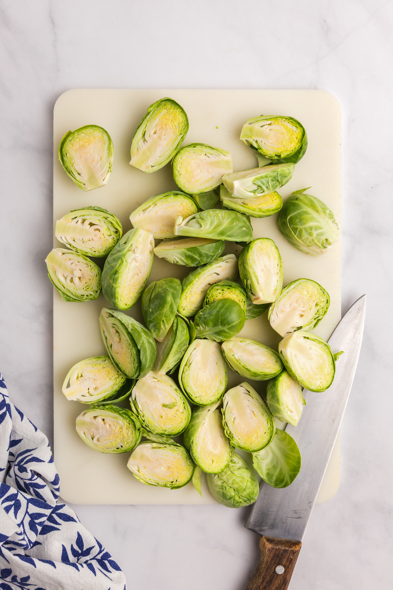 Fresh Brussels sprouts cut in half on a white cutting board with a knife placed to the side.