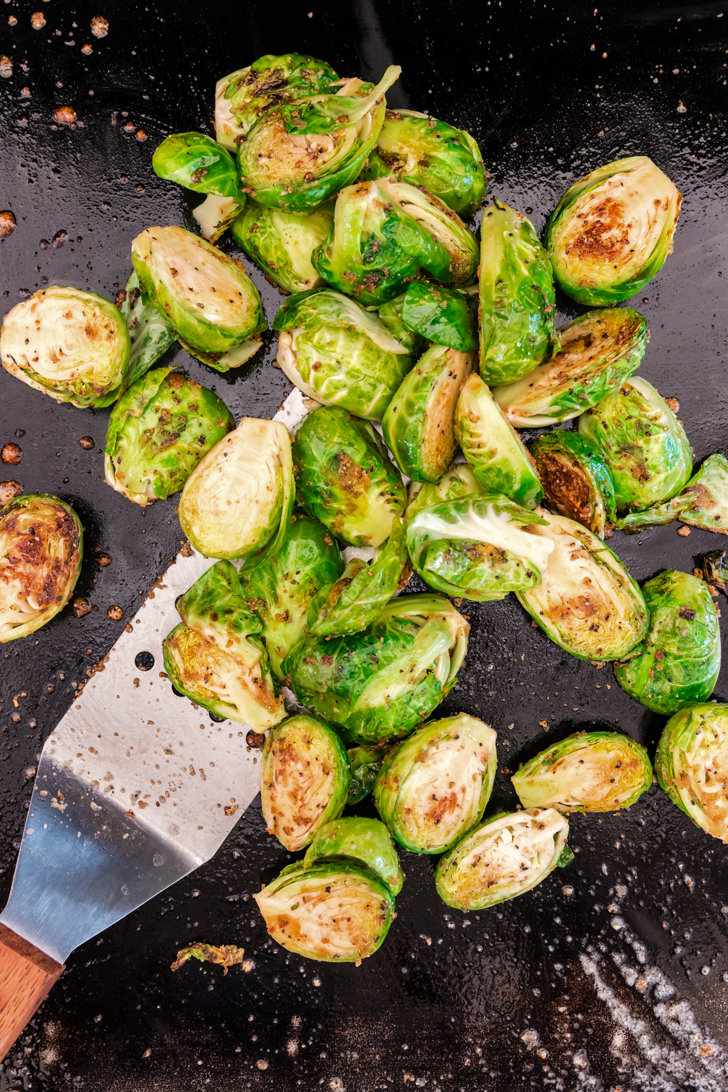Brussels sprouts cooking on a flat-top grill, starting to brown slightly with a spatula nearby.